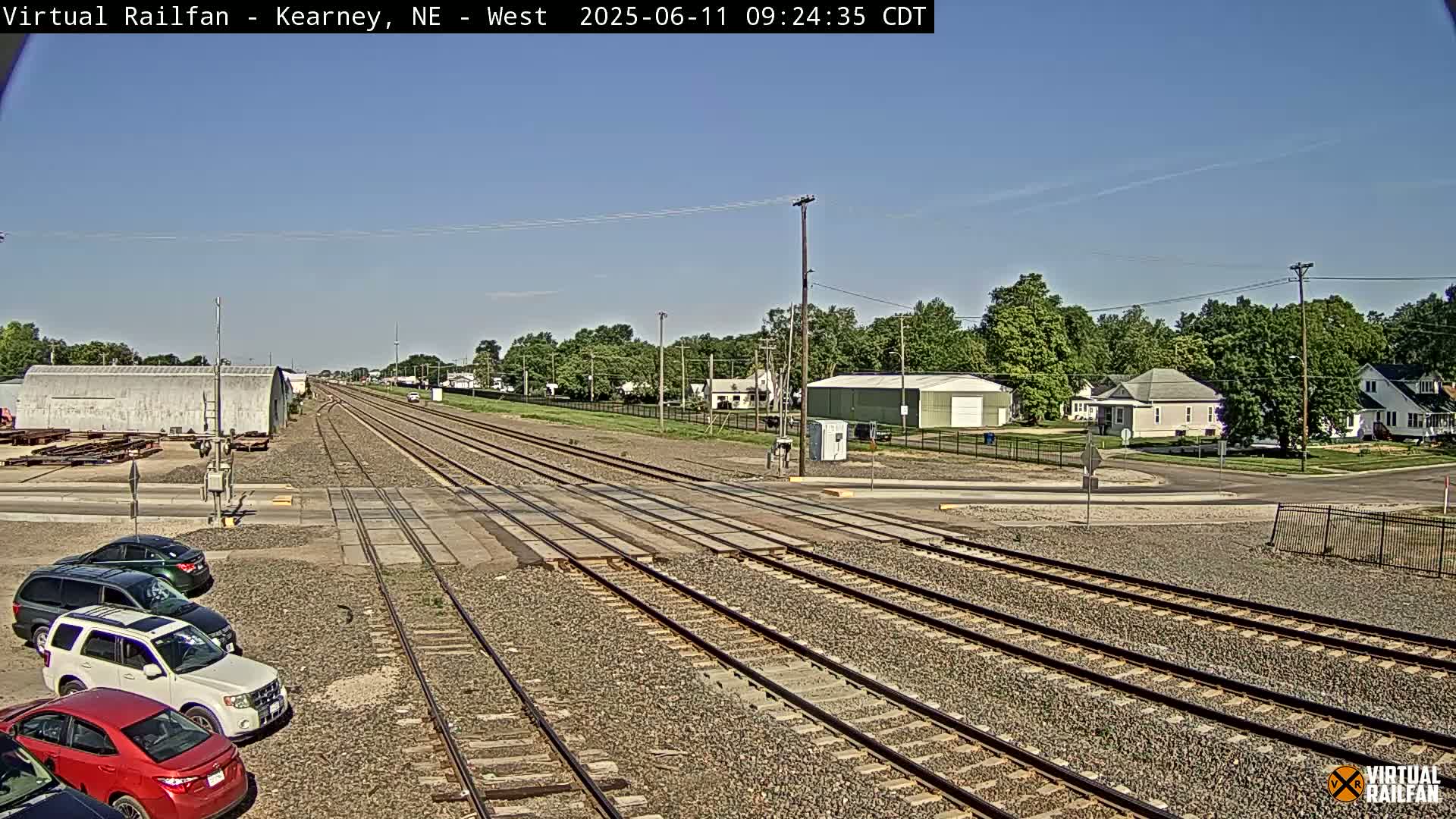 Multiple sets of train tracks intersect a road near a residential area under a clear, sunny sky.