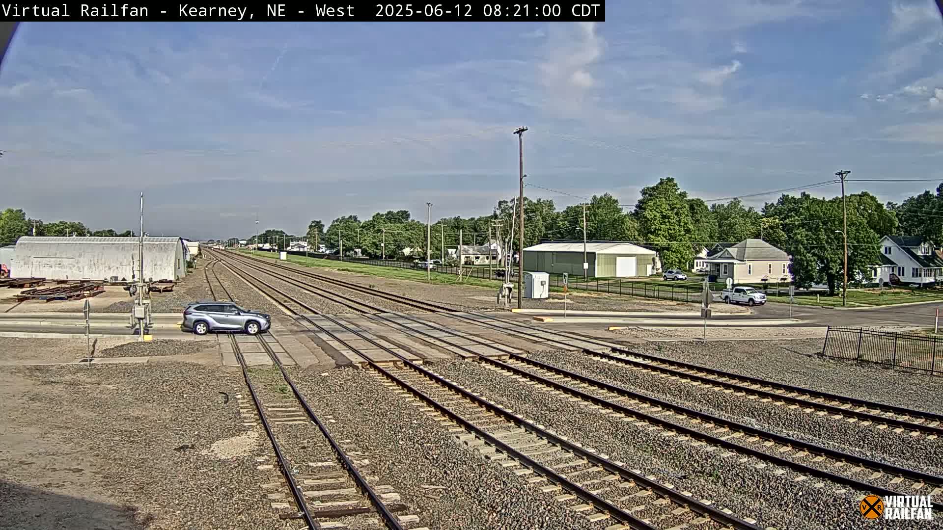 A silver SUV sits at a railroad crossing on a partly sunny day, with multiple train tracks extending into the distance past a small town with green trees and buildings.