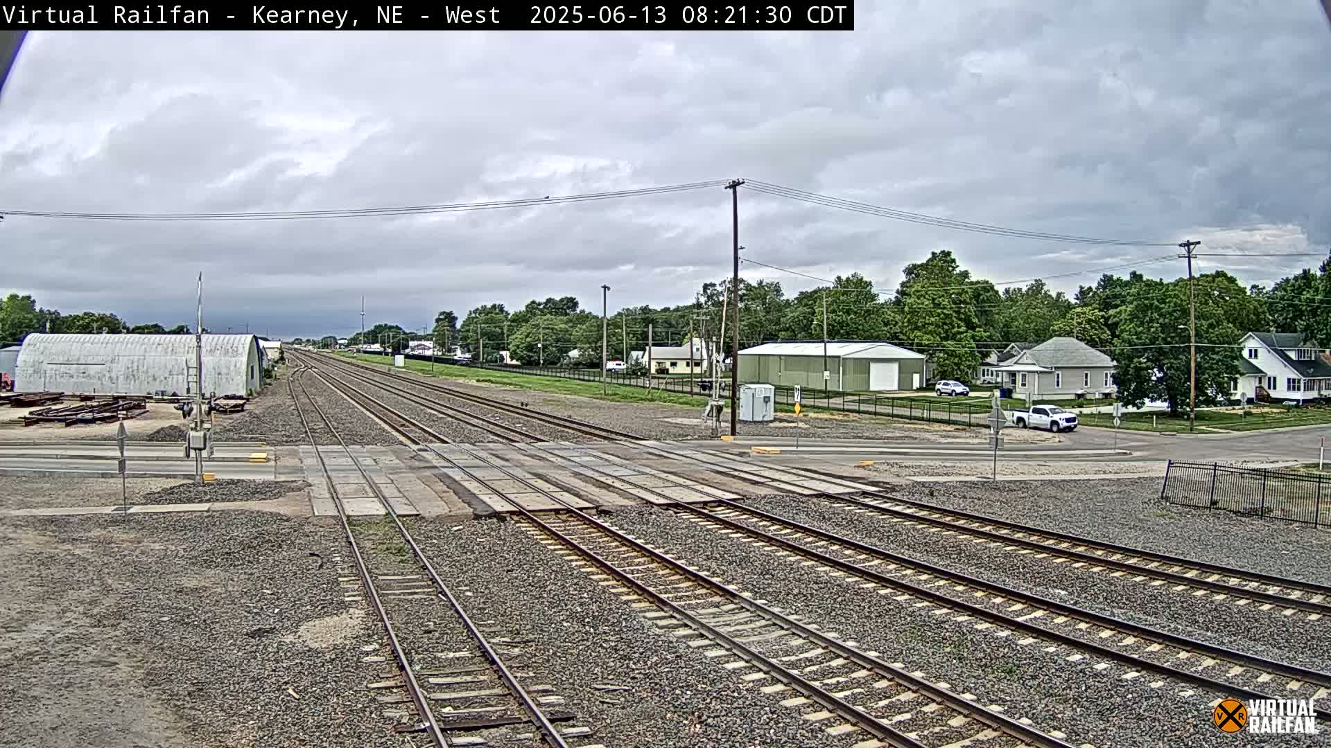 A railroad crossing in a small town under a cloudy sky shows multiple train tracks intersecting a road with a few buildings and vehicles visible in the background.