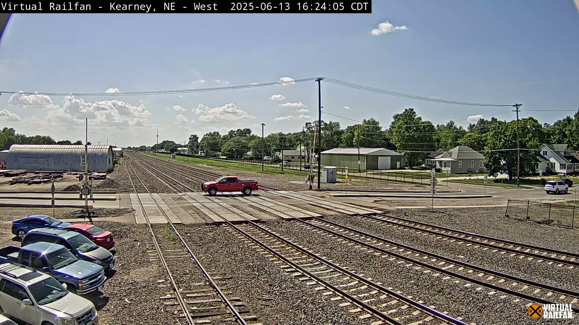 A red pickup truck is crossing a railroad crossing in a town on a partly cloudy day, with several cars parked nearby and buildings visible in the background.