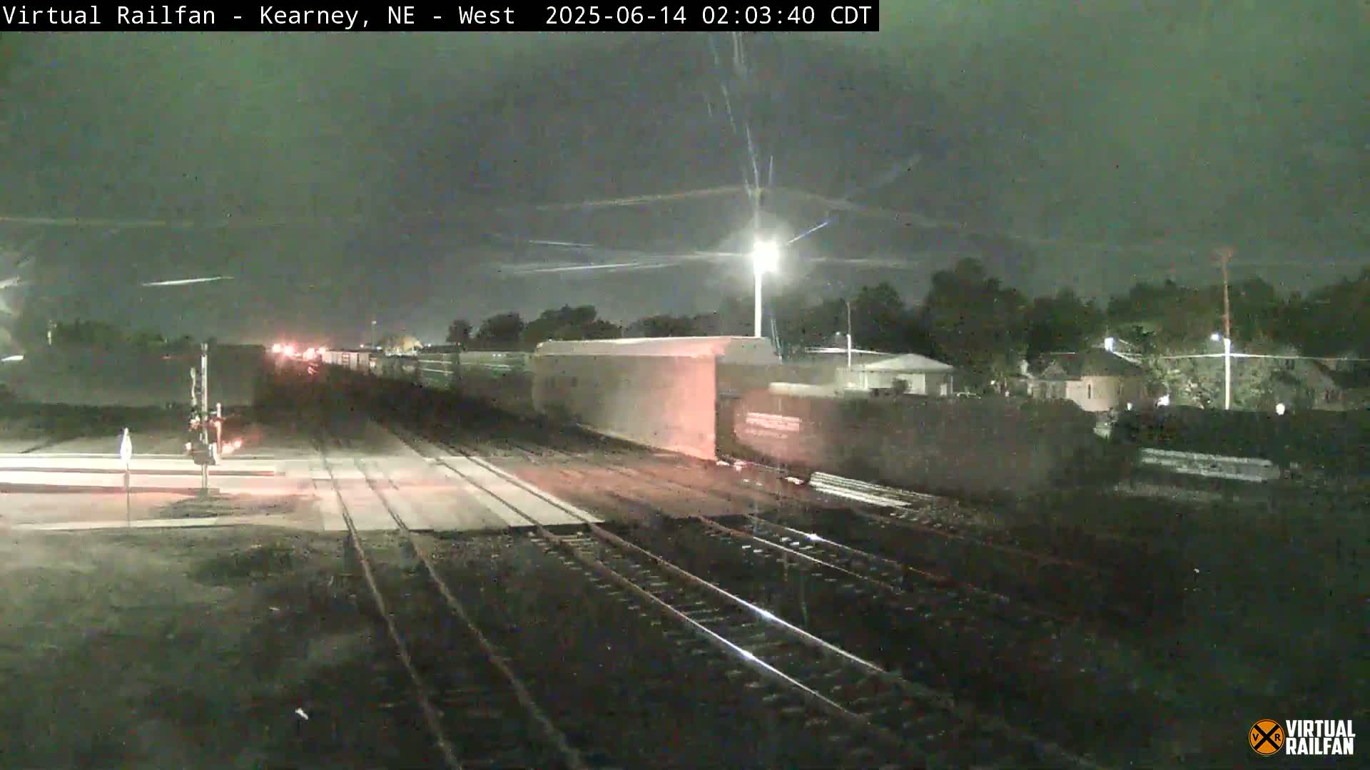 A nighttime view of a train passing through a railway crossing under a dark, possibly overcast sky.