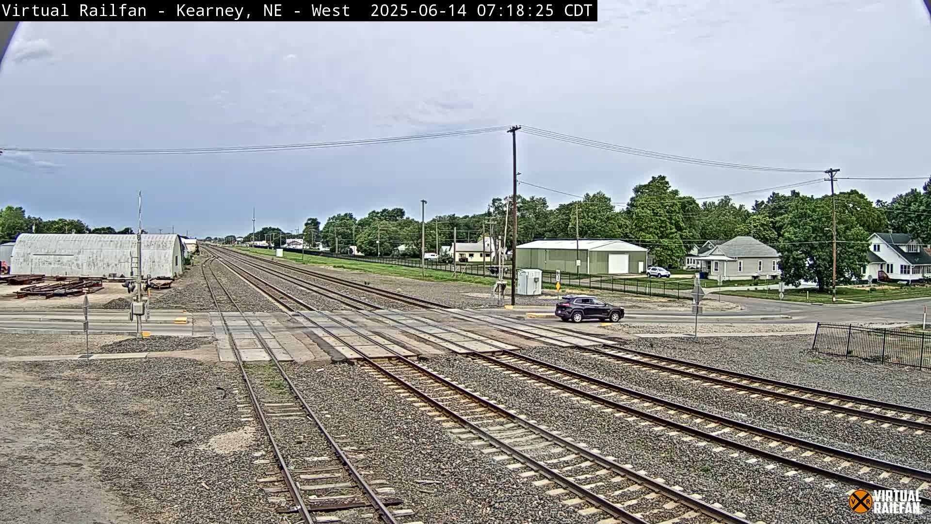 A mostly cloudy day shows multiple train tracks intersecting a road, with a dark-colored SUV stopped at the crossing and buildings and trees in the background.