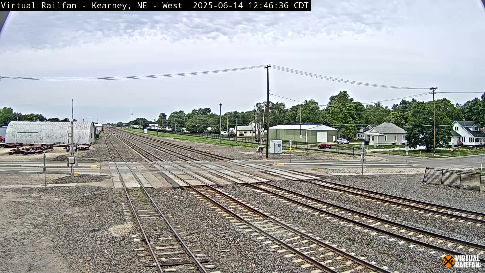 A railroad crossing in a small town on an overcast day shows multiple train tracks intersecting a road with buildings and trees in the background.