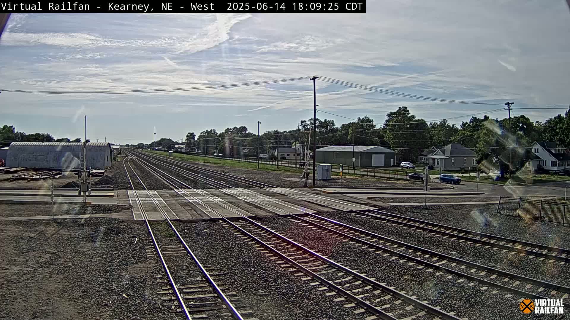 A railroad crossing in a small town on an overcast day shows multiple train tracks intersecting a road with buildings and trees in the background.
