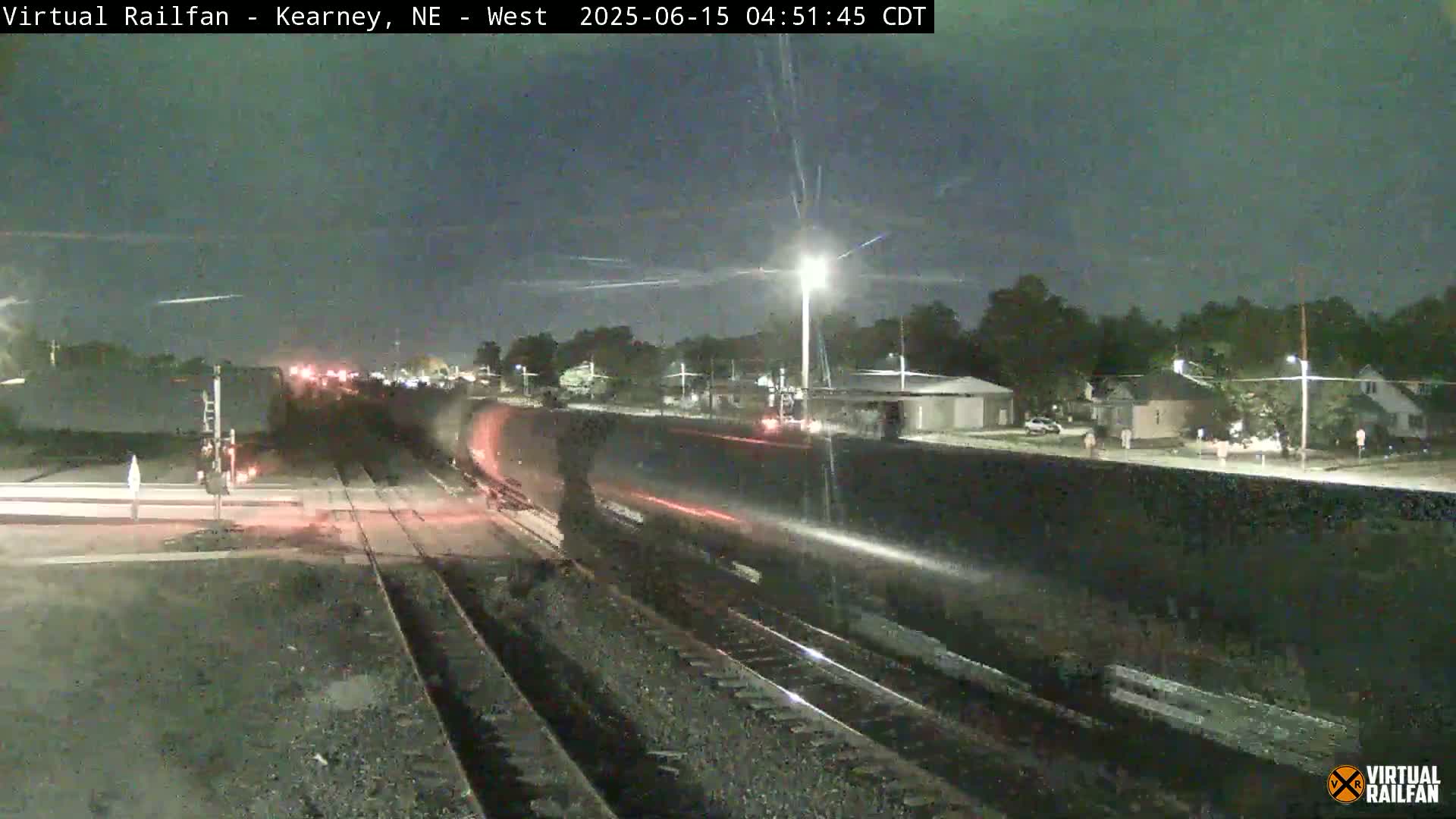 A long freight train passes under a nighttime sky illuminated by streetlights, near a road with houses and trees.