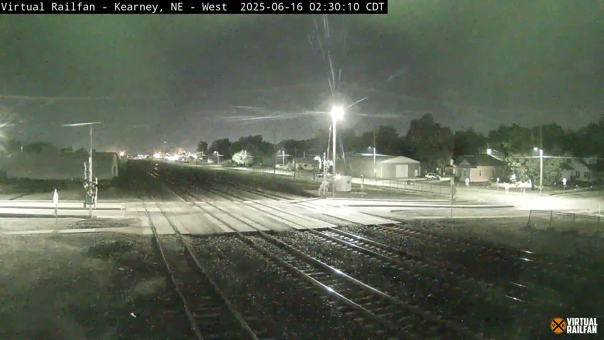 A nighttime view of a railroad crossing with a street intersection under a partly cloudy sky.