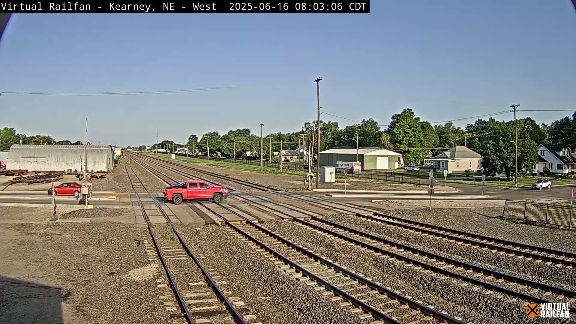 A red pickup truck and a red car are stopped at a railroad crossing on a sunny day, with railroad tracks extending into the distance past buildings and trees.