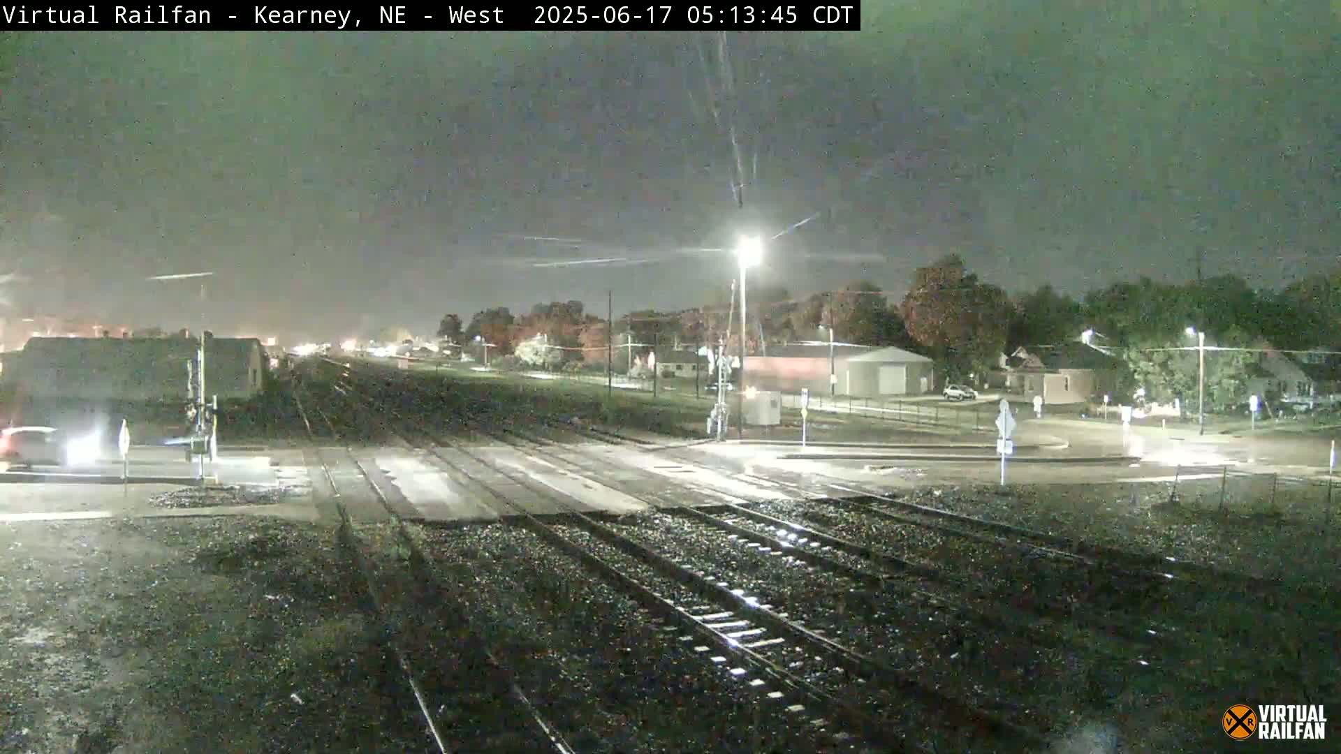 A nighttime view of a wet railroad crossing in a residential area, under a dark, possibly overcast sky.
