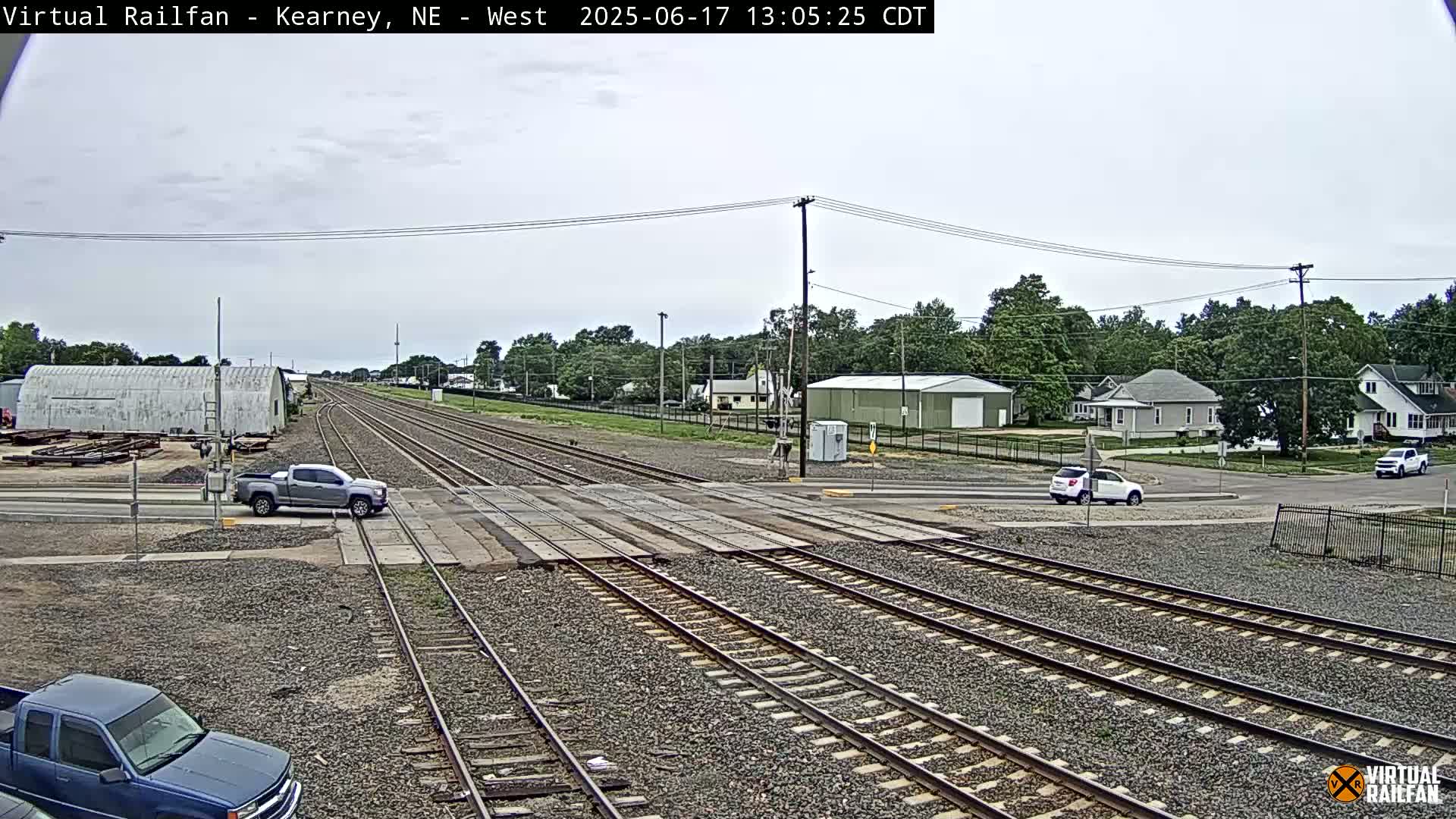 A railroad crossing in a small town on an overcast day shows multiple train tracks, a few vehicles at the crossing, and houses and buildings in the background.