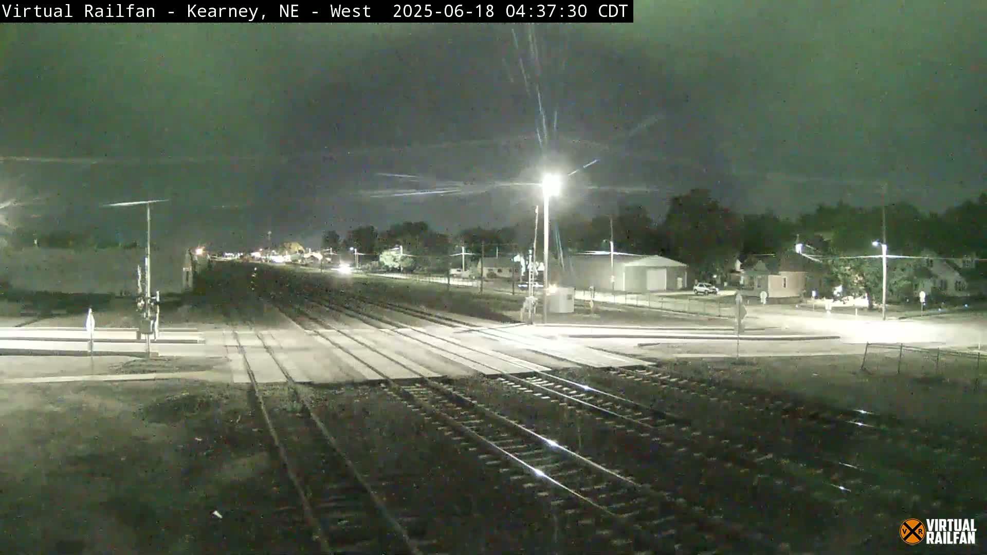 A nighttime view of railroad tracks intersecting a road, with sparsely lit buildings and trees visible in the background under a mostly clear sky.