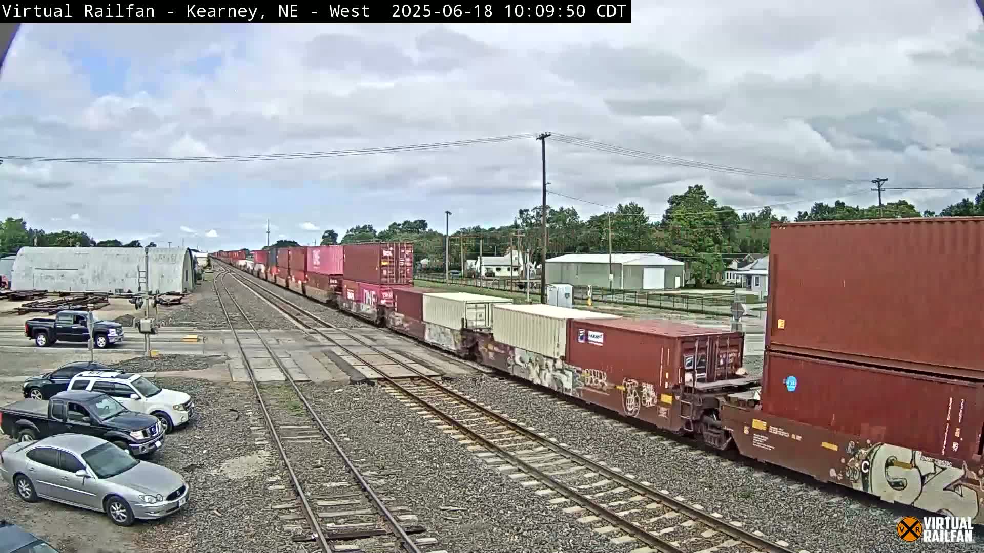A long freight train carrying numerous shipping containers passes through a railway crossing under a partly cloudy sky.