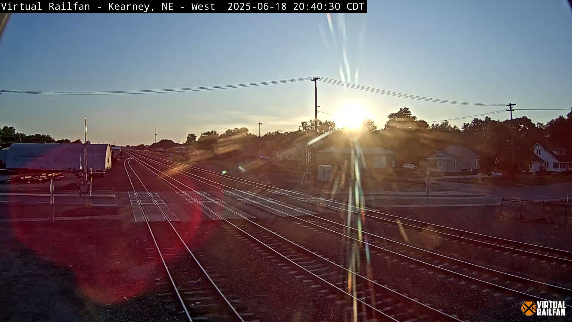 Multiple sets of railroad tracks extend into the distance toward a setting sun, with houses and trees visible in the background under a clear sky.