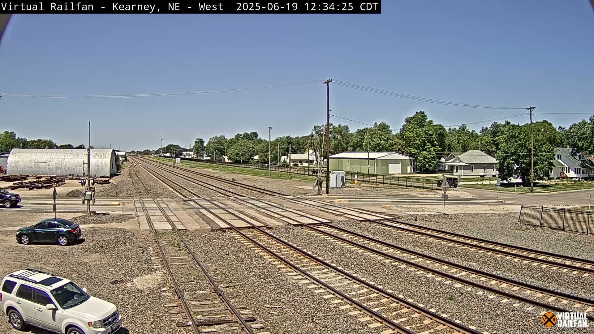 A railroad crossing in a residential area on a sunny day, with two cars visible near the tracks and several buildings in the background.
