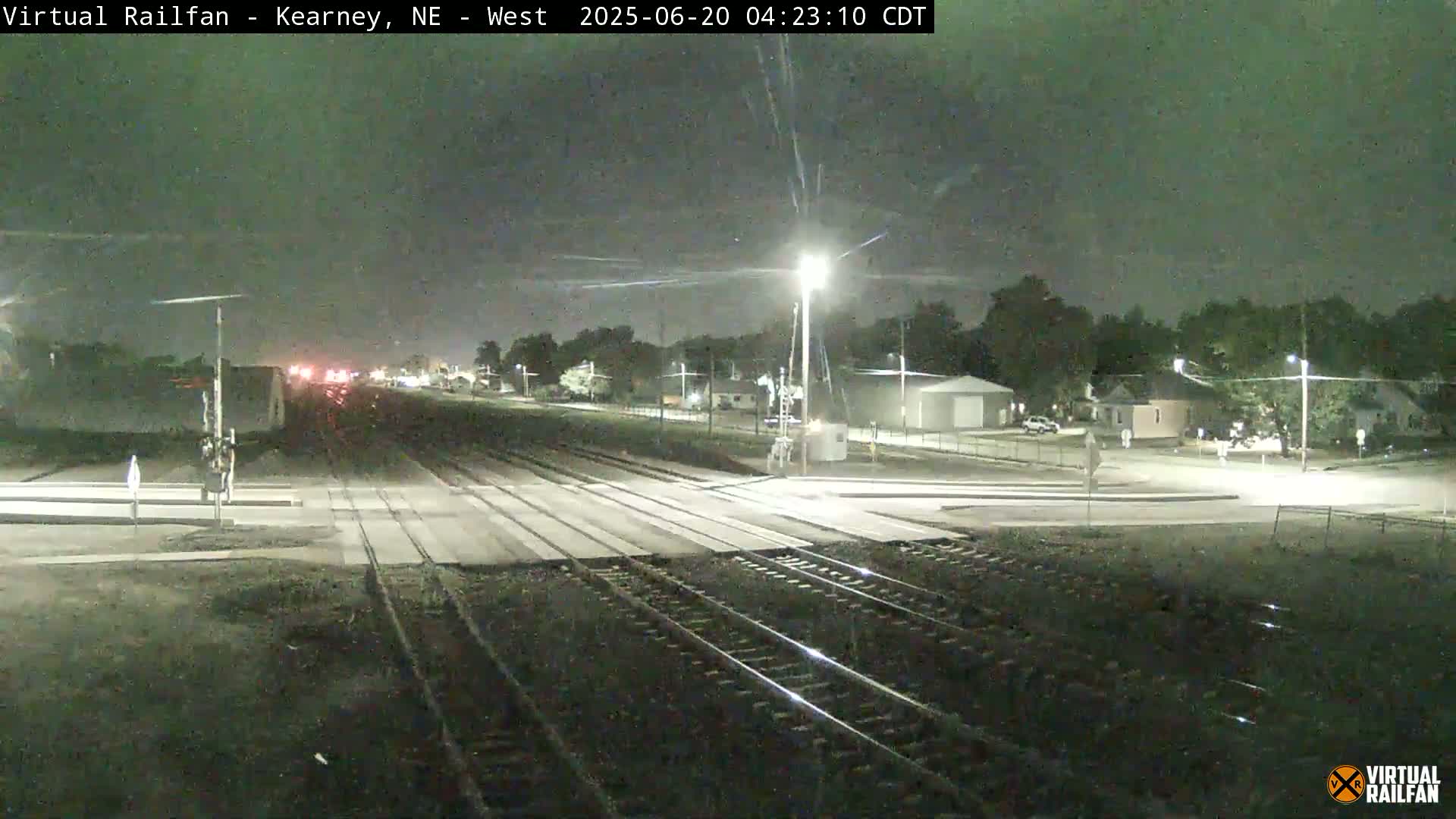A nighttime view of railroad tracks intersecting with a road, under a mostly clear sky, with a few houses and streetlights visible in the distance.