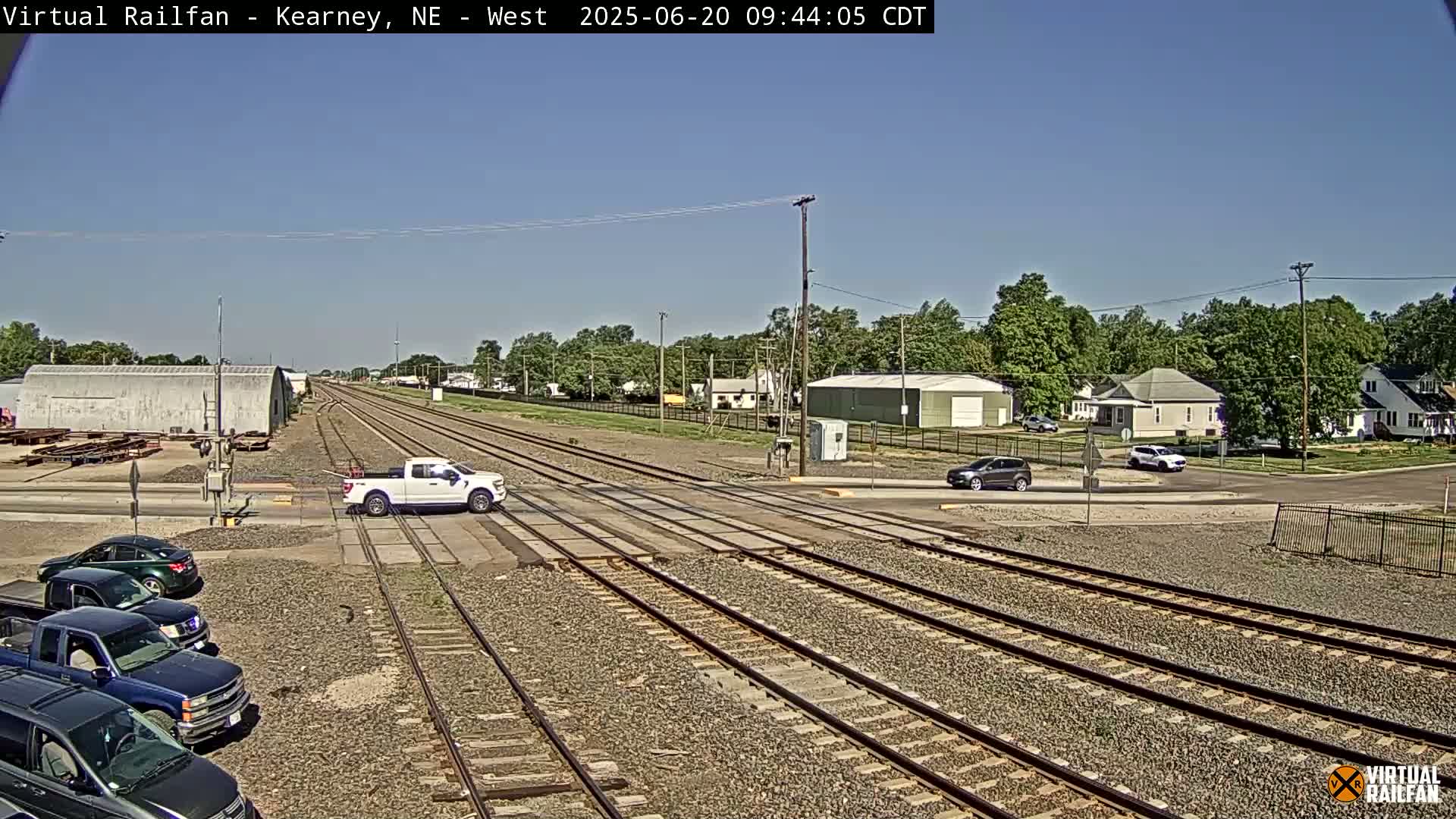A sunny day shows a railroad crossing in a small town with several cars at the intersection and parked nearby, and a white pickup truck crossing the tracks.