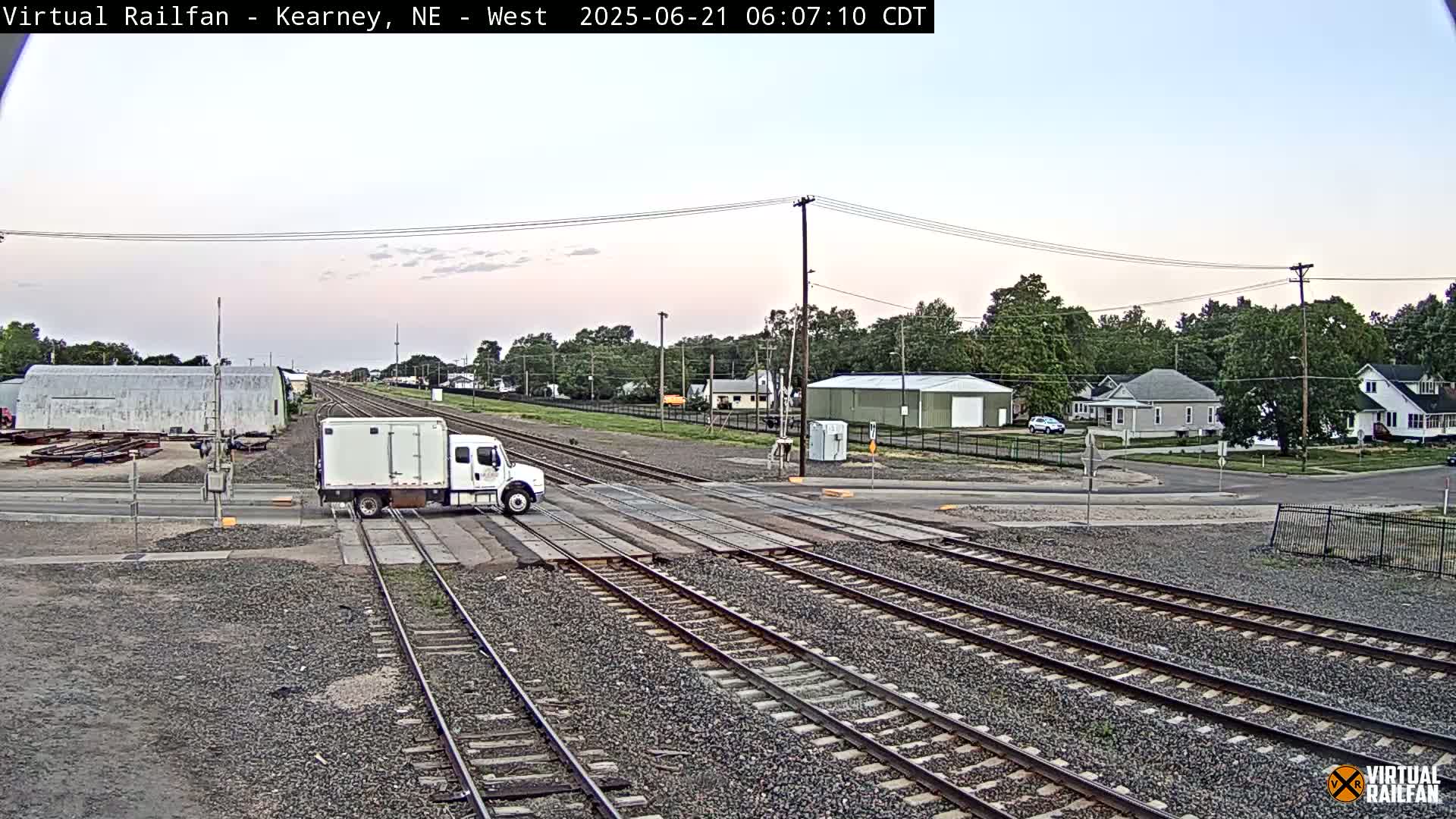 A white box truck is crossing a railroad crossing in a residential area under a partly cloudy sky.