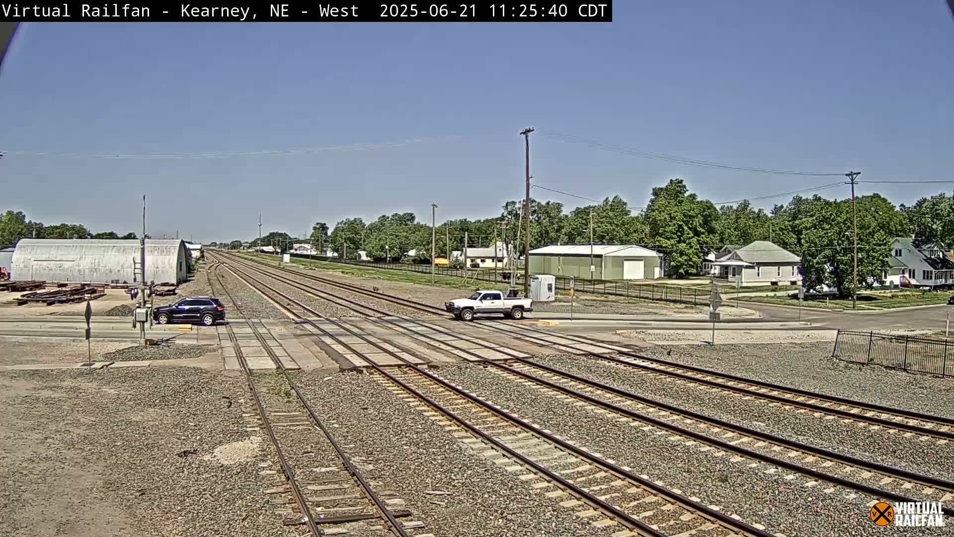 A sunny day reveals a railroad crossing with multiple tracks, a few vehicles, and residential buildings in the background.
