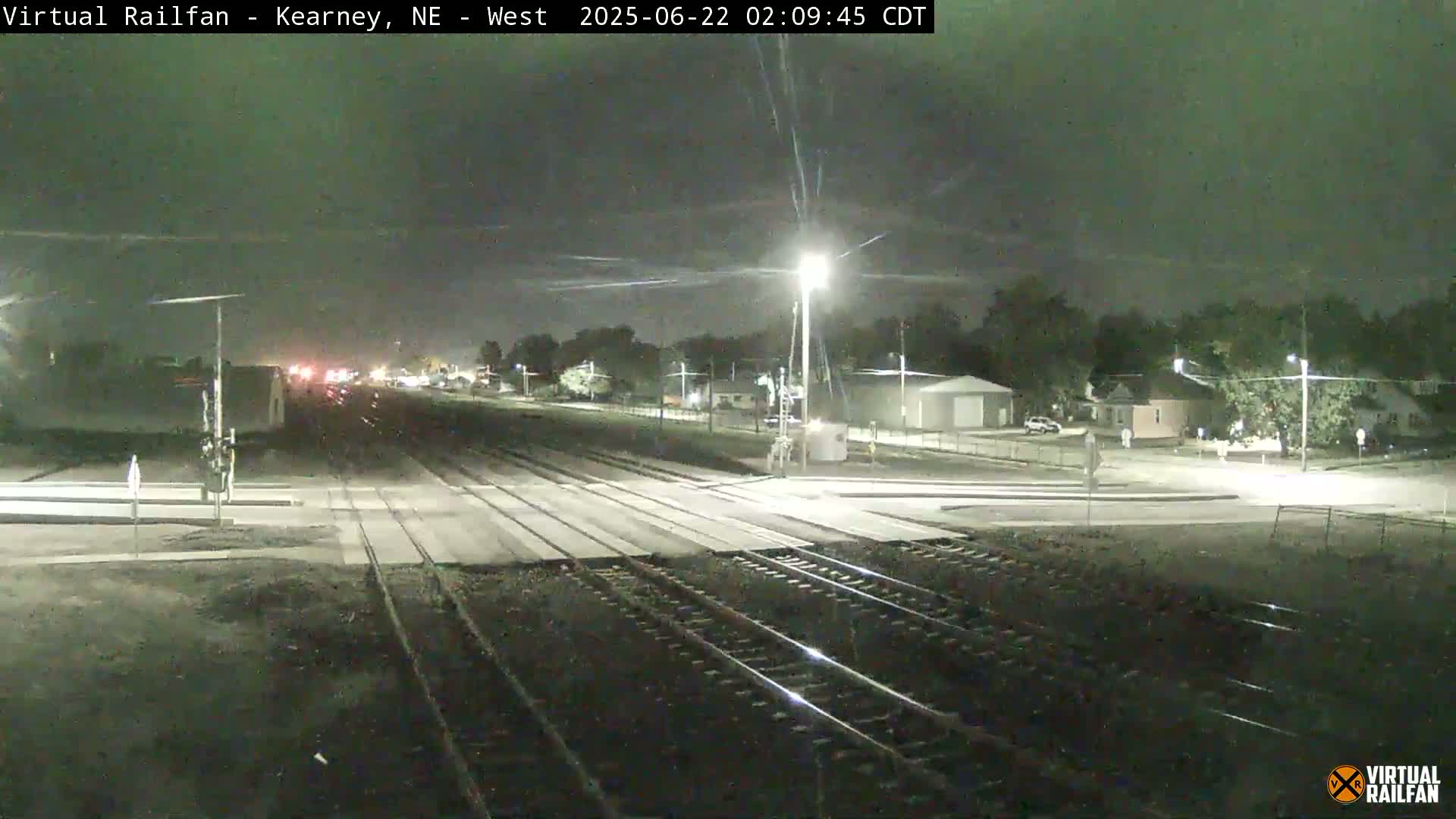 A nighttime view of railroad tracks intersecting a road, with a few houses and streetlights visible in the background under a dark sky.