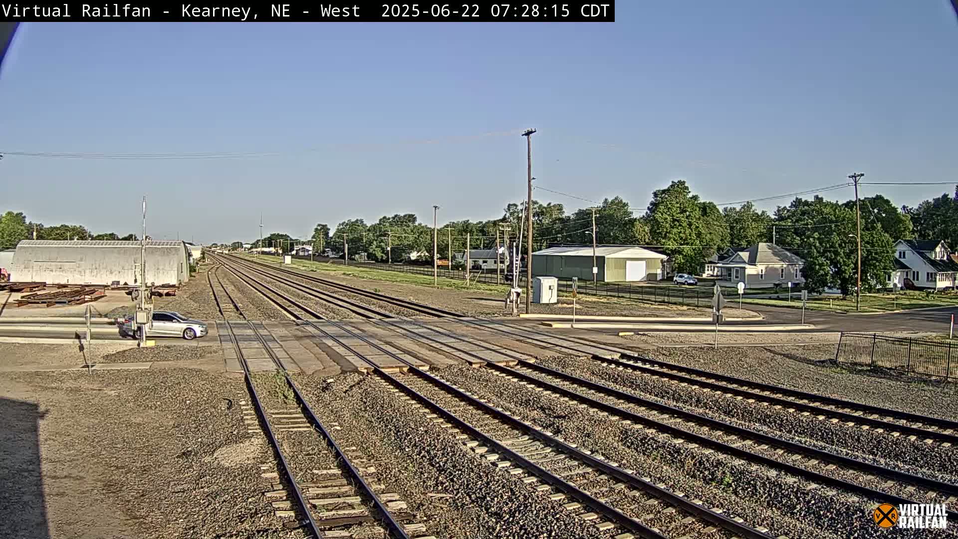 A sunny day shows multiple train tracks intersecting with a road, a car waiting at the crossing, and a residential area in the background.
