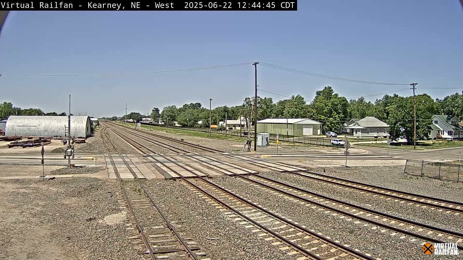 A railroad crossing intersects a road in a town on a sunny day, with trees and houses visible in the background.