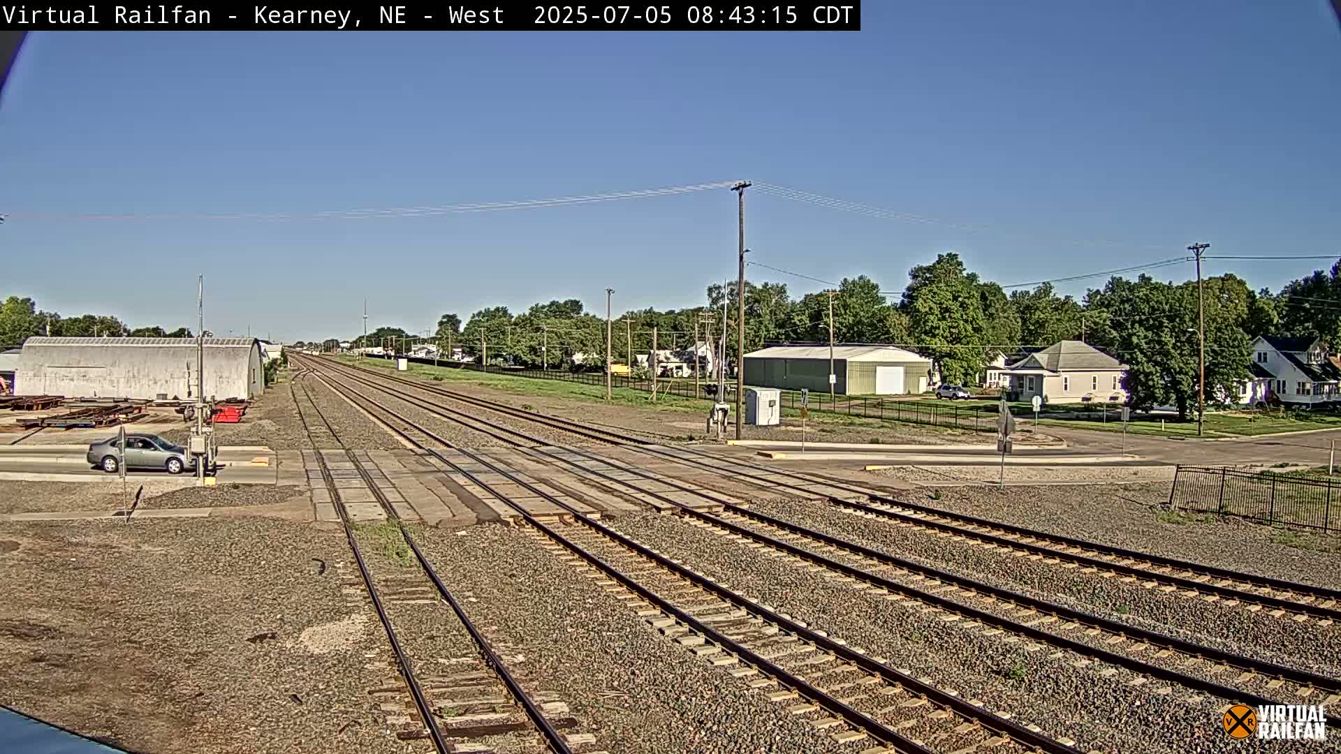 A long view of several train tracks intersecting with a road in front of residential buildings on a clear, sunny day.
