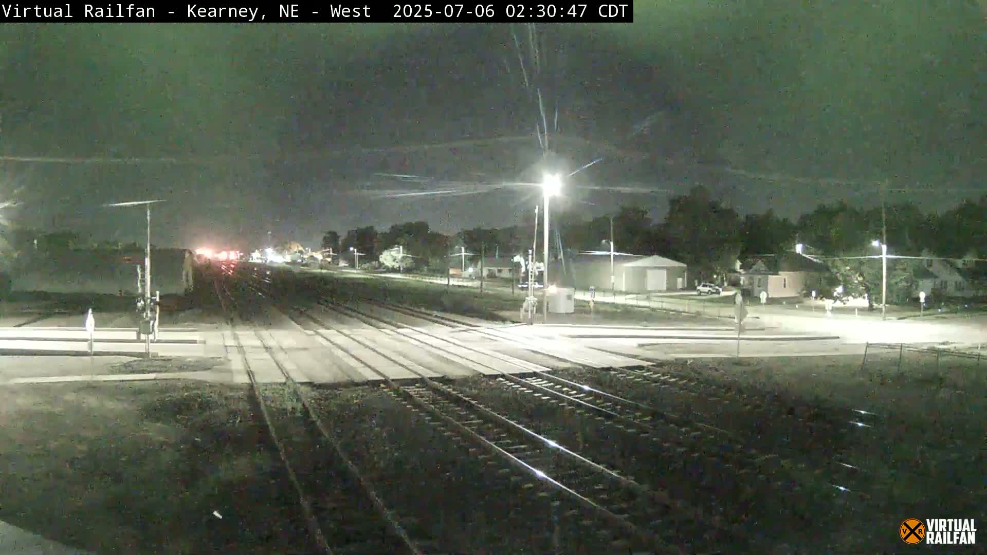 A nighttime view of railroad tracks intersecting a road, with a few houses and streetlights visible in the background under a clear sky.