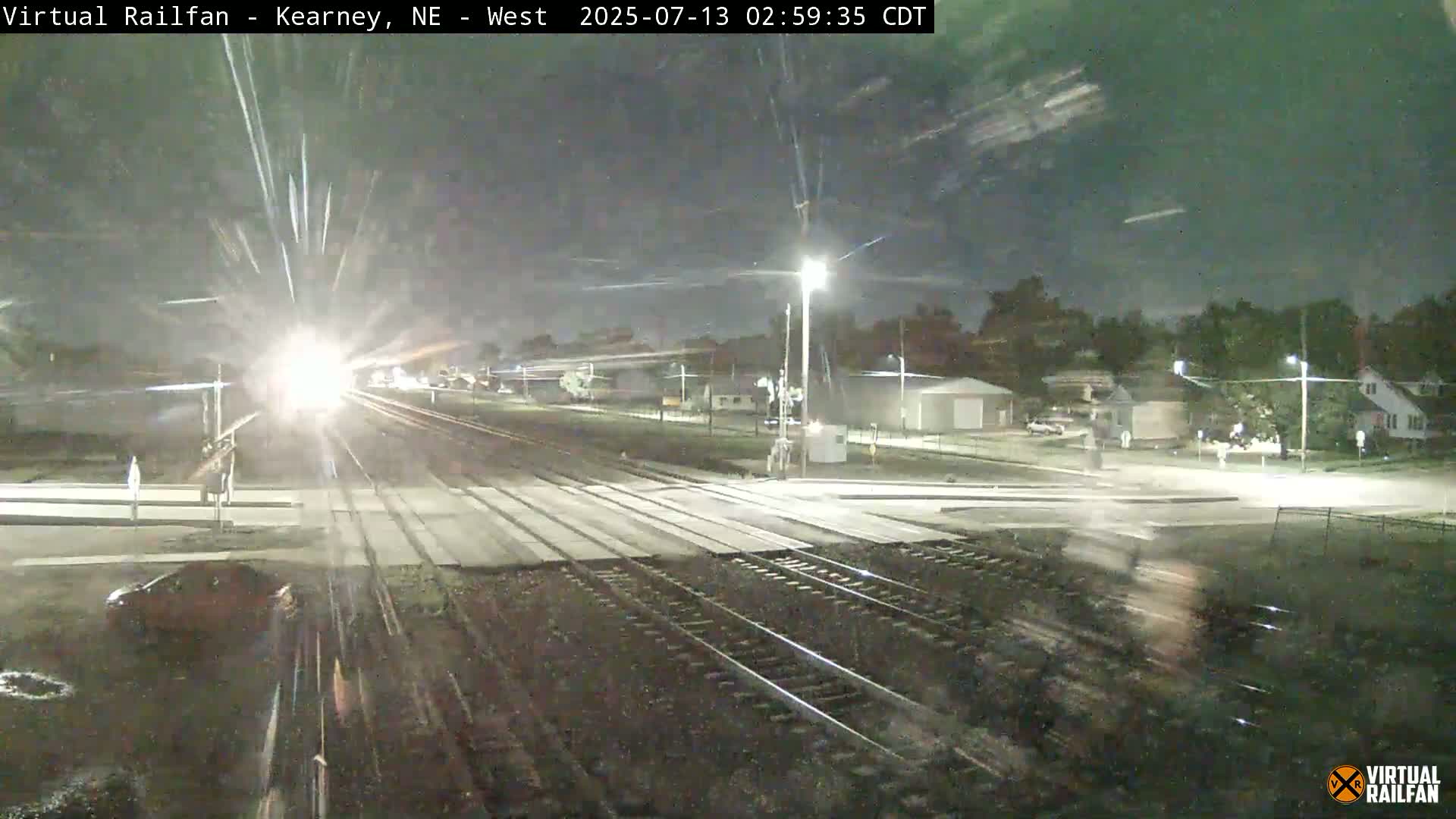 A brightly lit train passes a nighttime road crossing near some houses under a clear sky.