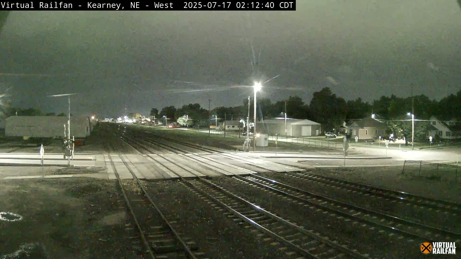 A nighttime view of railroad tracks intersecting a road, with a residential area visible in the background under a dark, overcast sky.