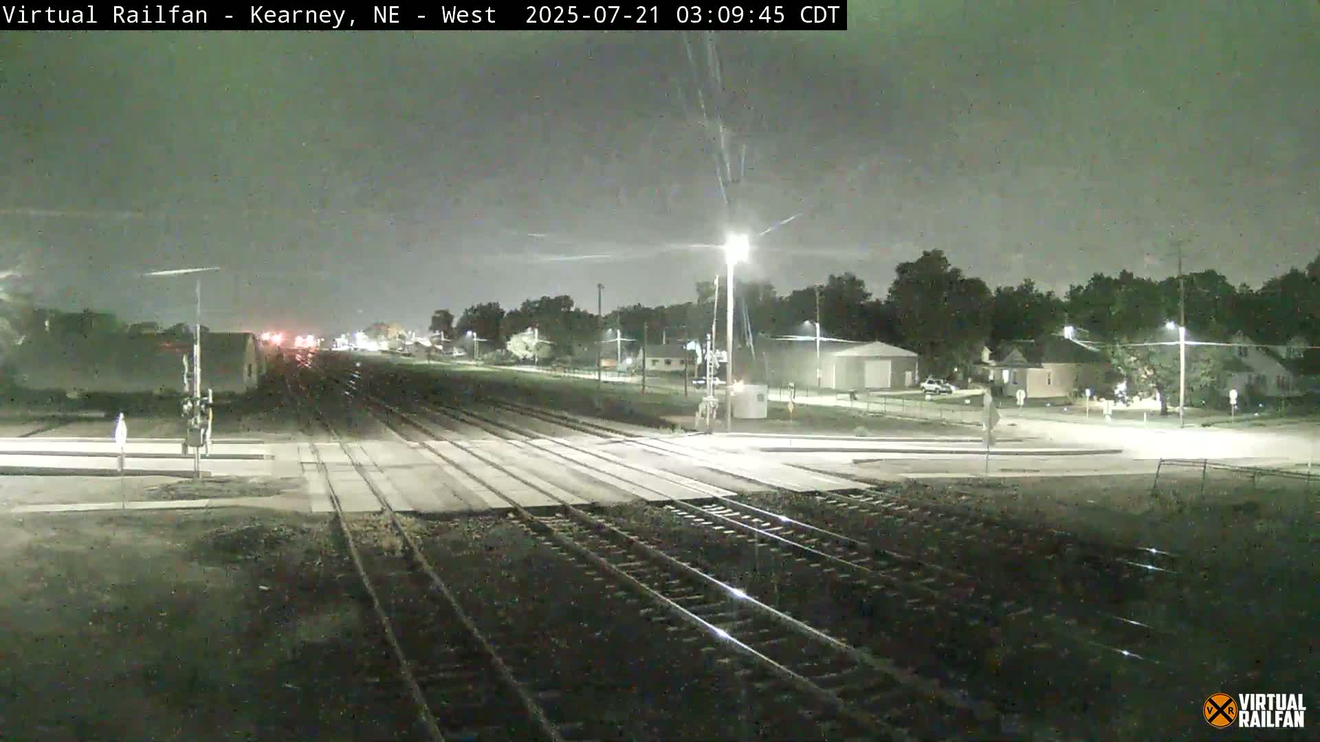A nighttime view of a railroad crossing with streetlights illuminating a quiet residential area under a cloudy sky.