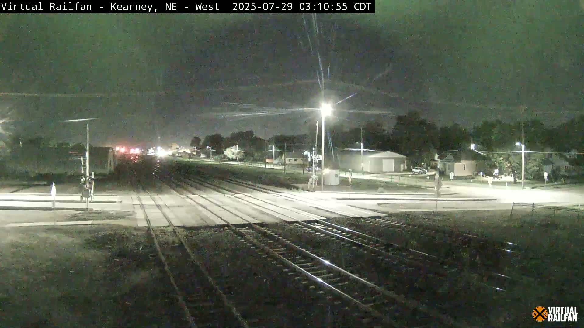 A nighttime view of a railroad crossing with train tracks extending into the distance, passing through a residential area under a dark sky.