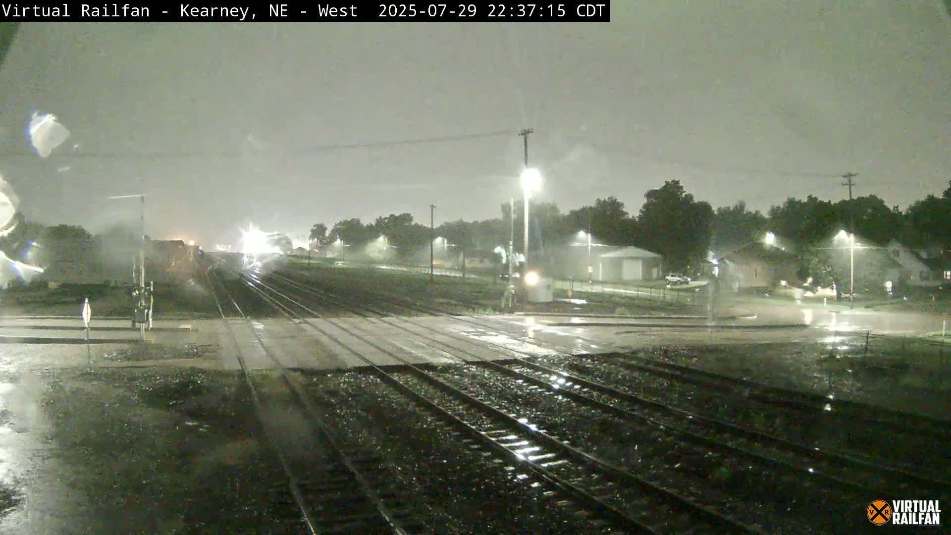 A nighttime view of a railroad crossing with train tracks extending into the distance, passing through a residential area under a dark sky.