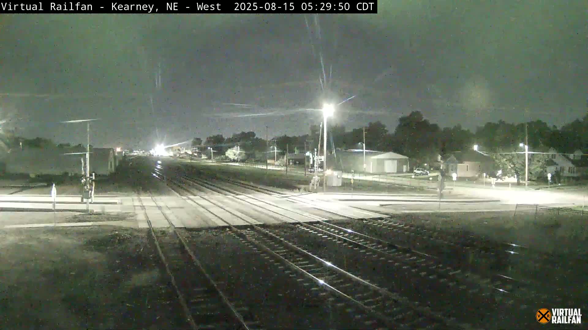A nighttime view of a railroad crossing with train tracks extending into the distance, passing through a residential area under a dark sky.