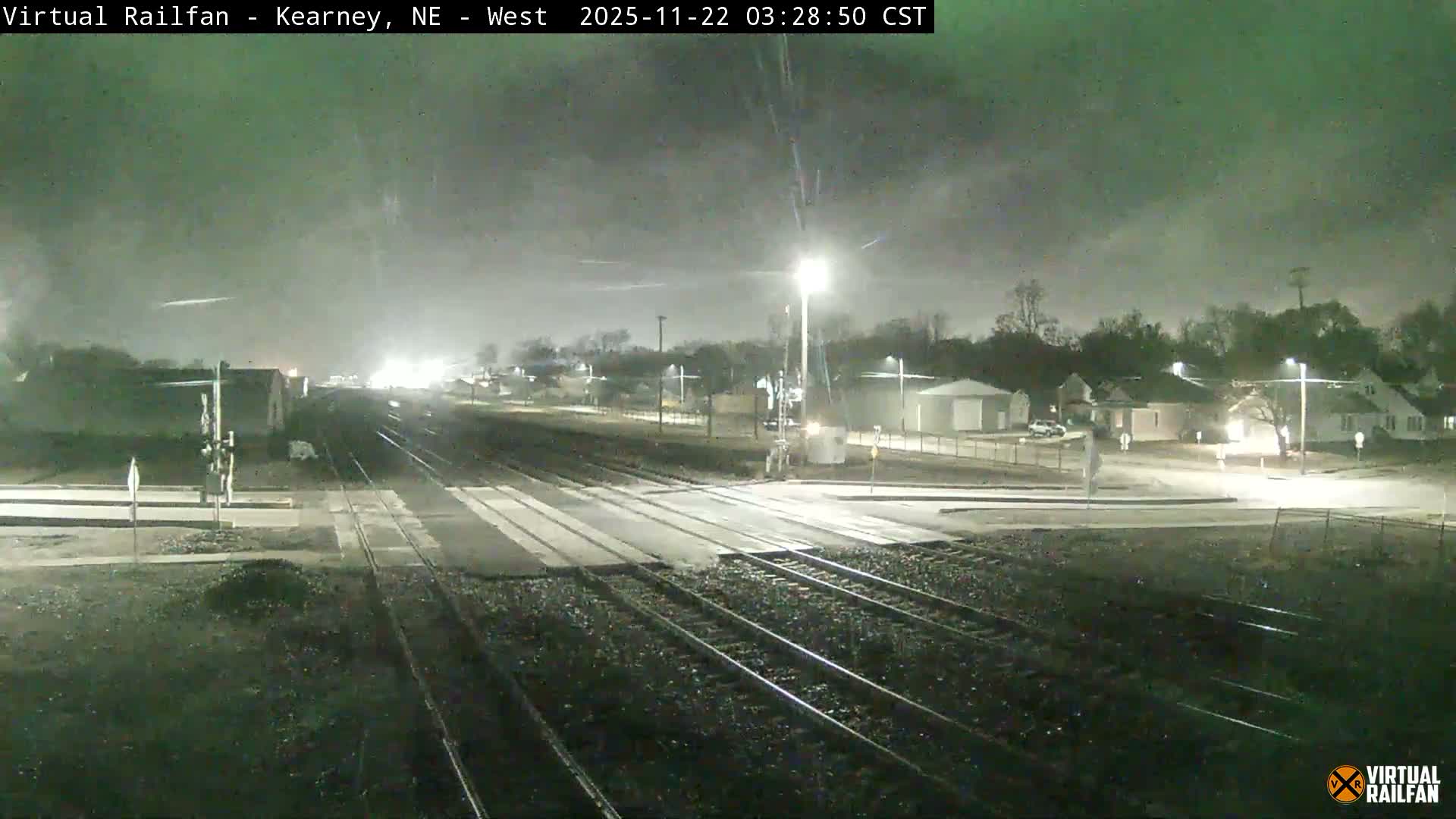 Multiple train tracks converge towards the horizon as they cross a road at night, illuminated by streetlights under an overcast sky, with residential buildings and sparse trees in the background.