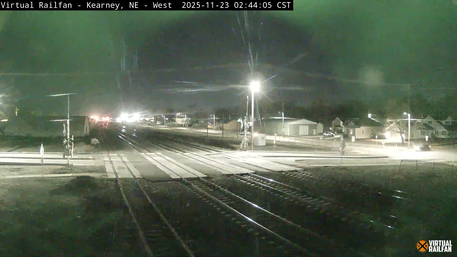 A dark outdoor scene at night shows multiple railroad tracks crossing a road with a level crossing, illuminated by streetlights and distant vehicle lights under a faintly green sky that appears to be experiencing light precipitation.