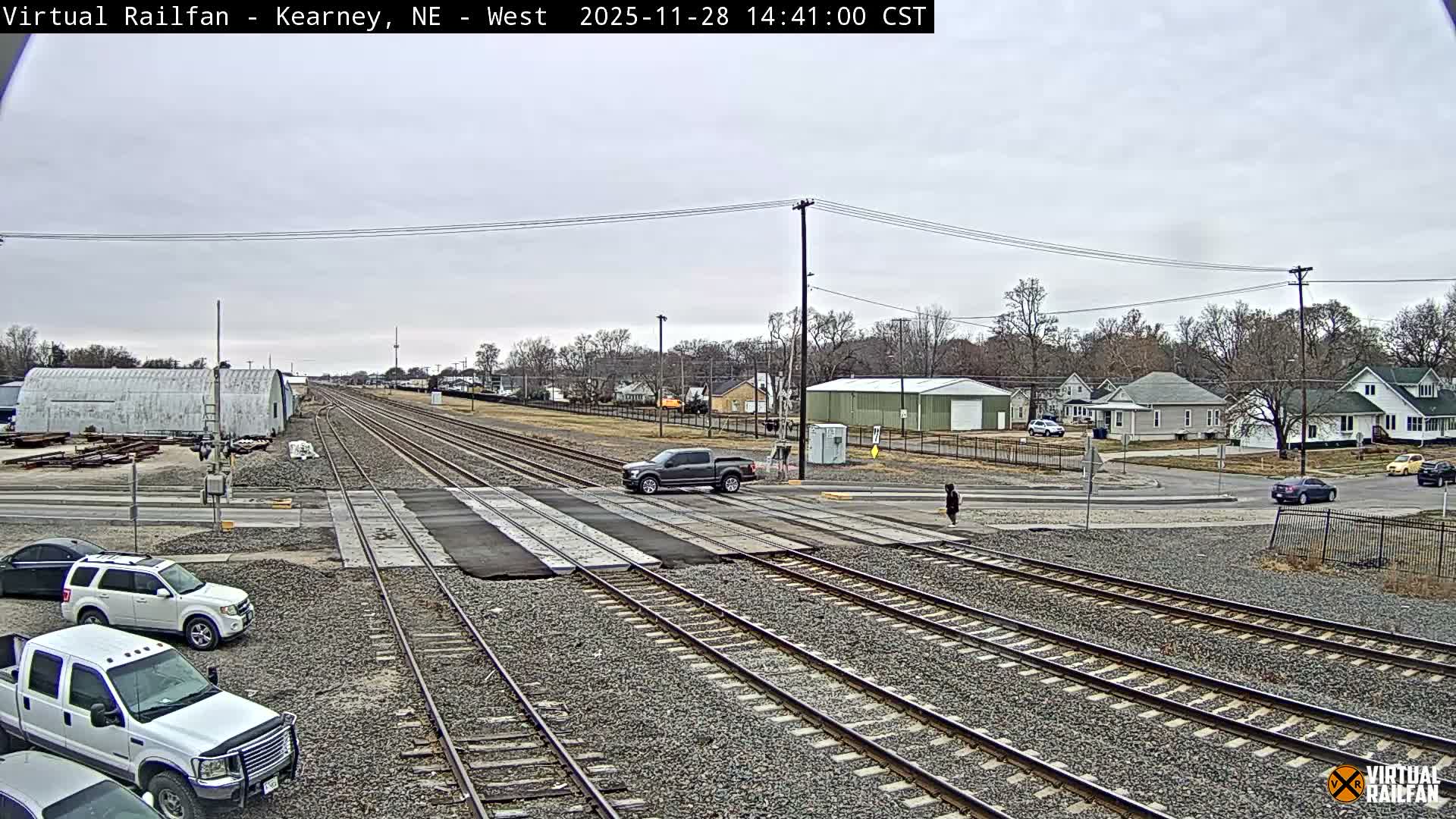 Multiple railroad tracks dominate the foreground and middle ground, with a pickup truck crossing them, while several cars are parked nearby and a person stands by the crossing, all under an overcast sky with residential and industrial buildings in the background.