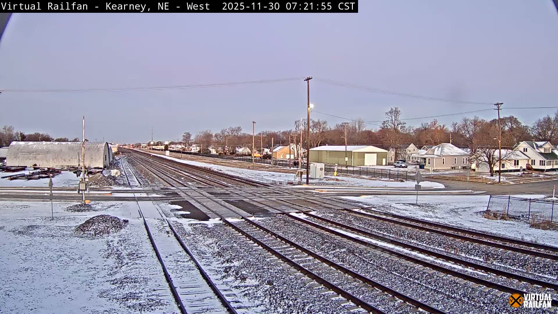 Multiple railroad tracks dominate the foreground and middle ground, with a pickup truck crossing them, while several cars are parked nearby and a person stands by the crossing, all under an overcast sky with residential and industrial buildings in the background.
