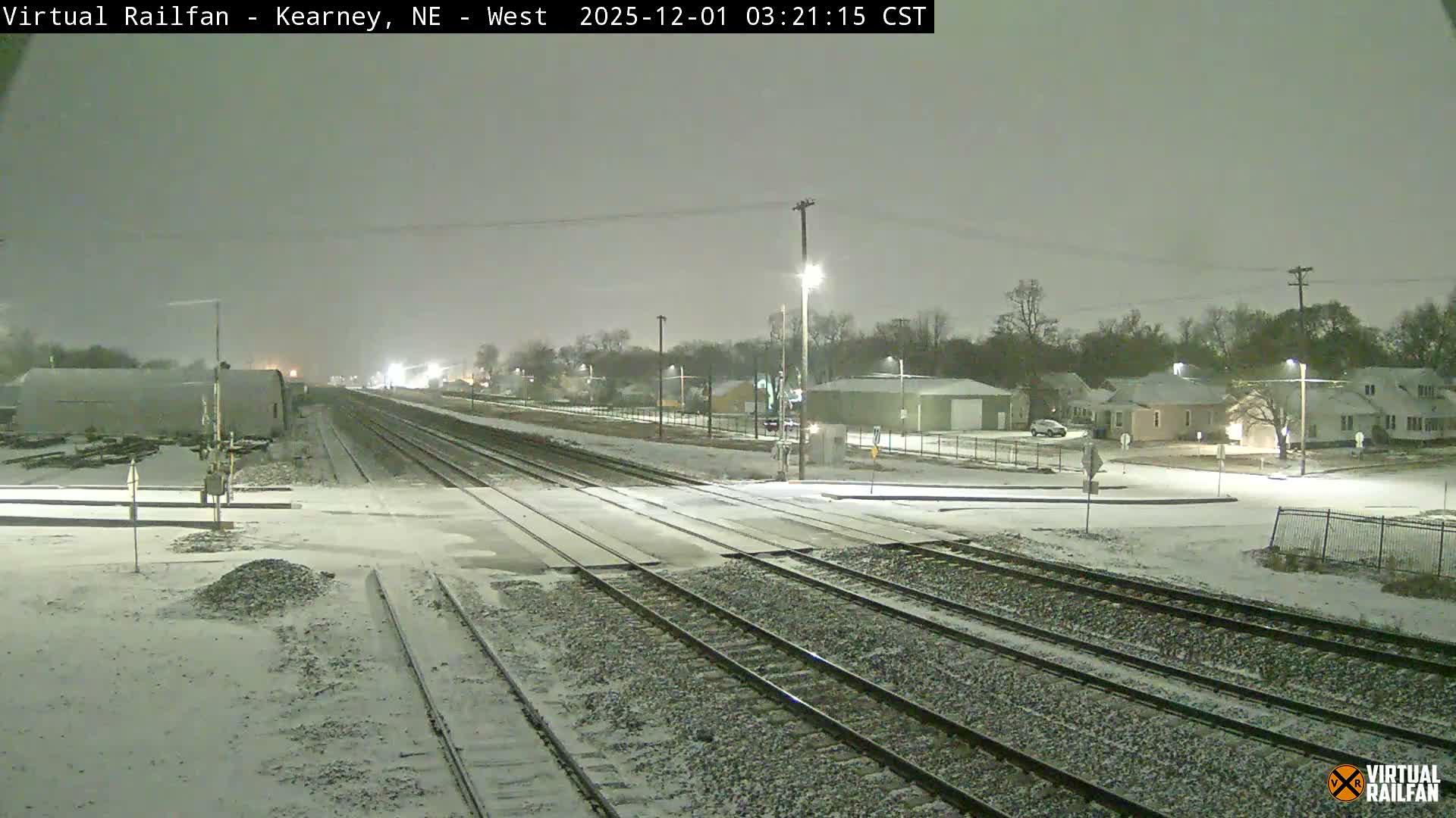 The image shows a snowy night scene with multiple railroad tracks extending into the distance, flanked by industrial buildings on the left and residential houses and a street illuminated by bright streetlights on the right.