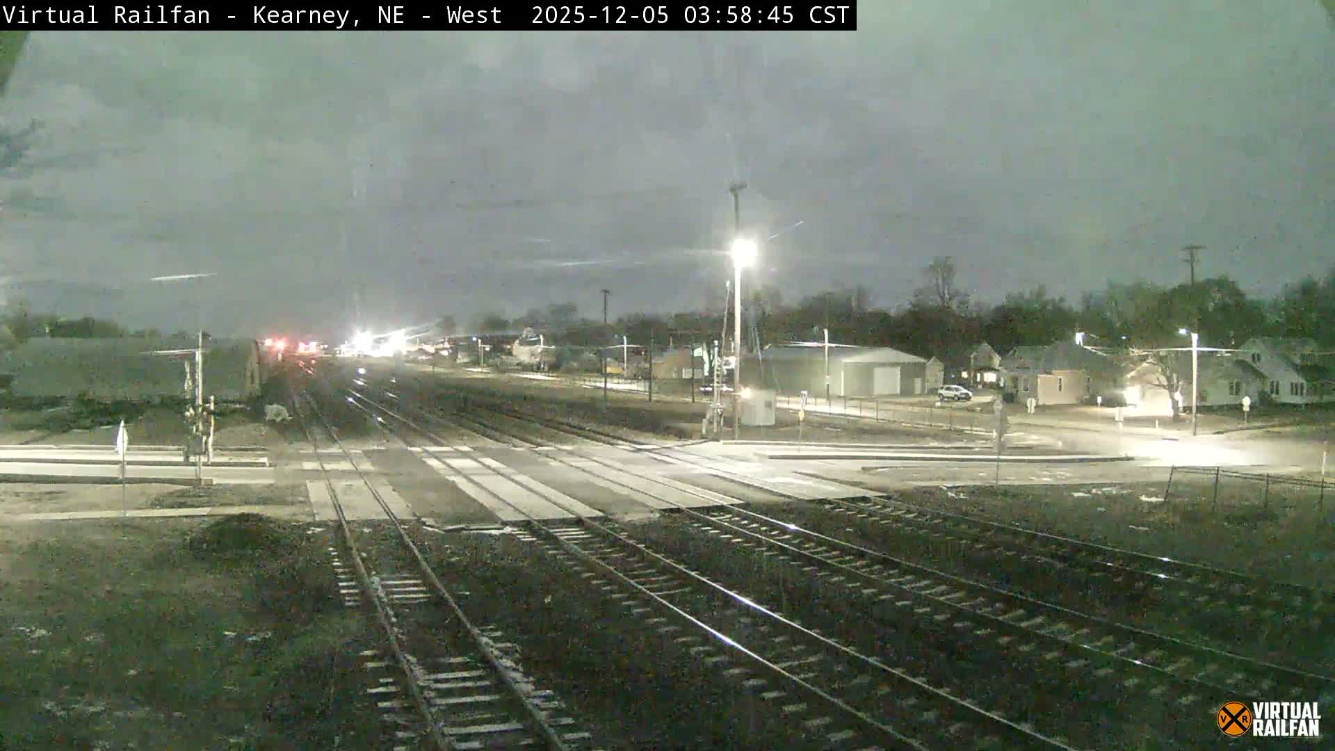 A multi-track railway crossing is illuminated by bright streetlights and the headlights of a distant train under a dark, overcast night sky, with residential houses and a white industrial building visible in the background.