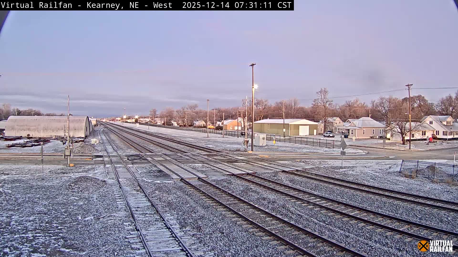 A multi-track railway crossing is illuminated by bright streetlights and the headlights of a distant train under a dark, overcast night sky, with residential houses and a white industrial building visible in the background.