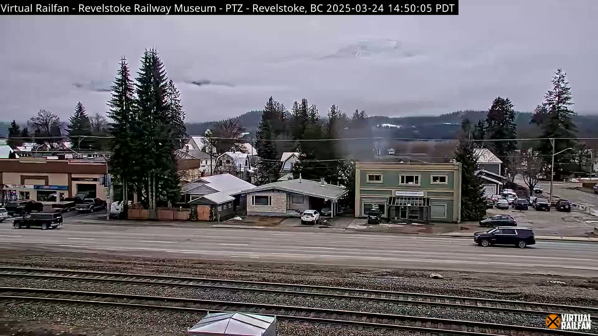 A cloudy day reveals a town with a mix of buildings and cars, situated alongside railroad tracks, with mountains partially obscured by clouds in the background.