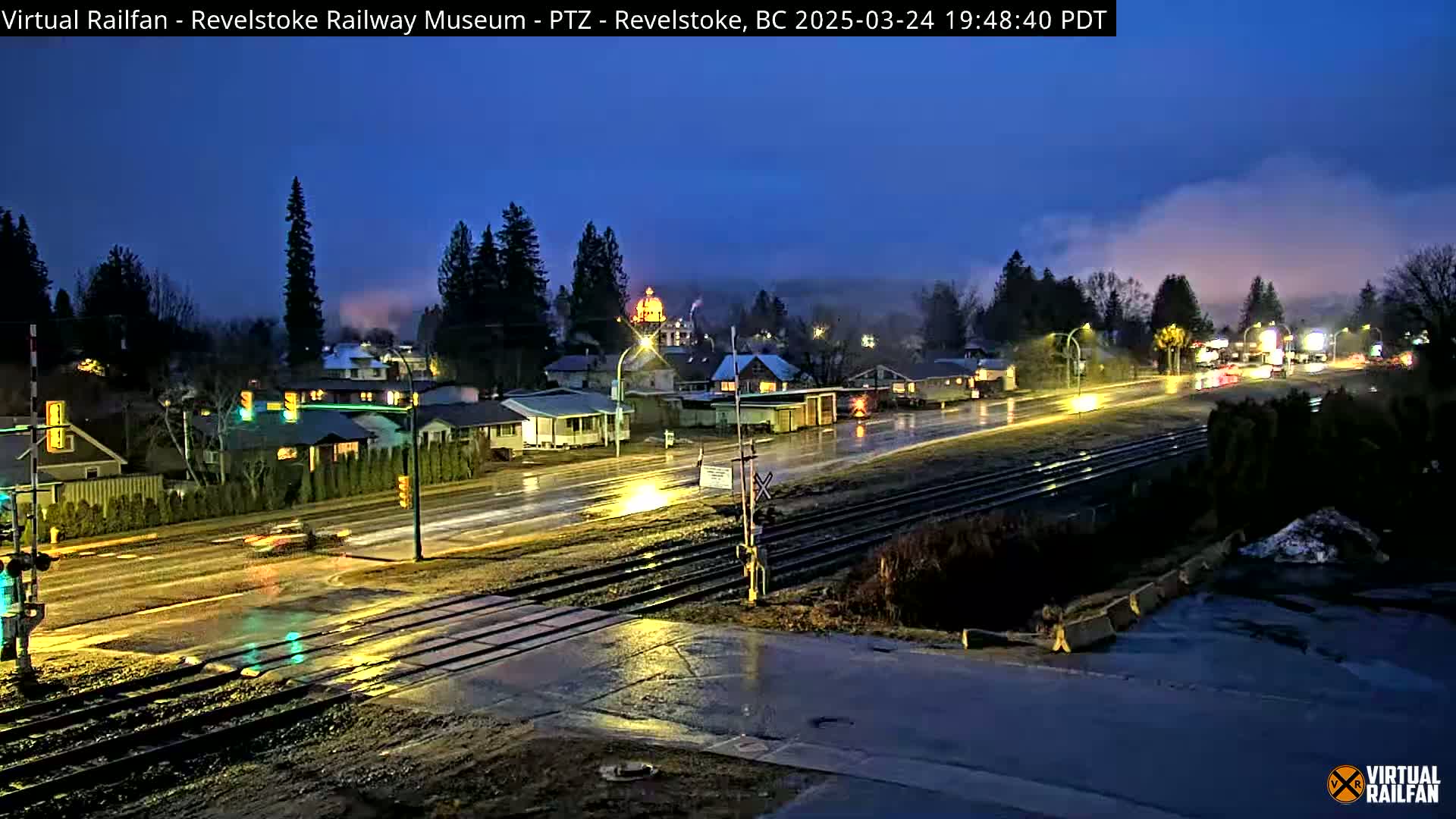 A wet, nighttime view of a town with a railway track running through it, under a mostly cloudy sky.