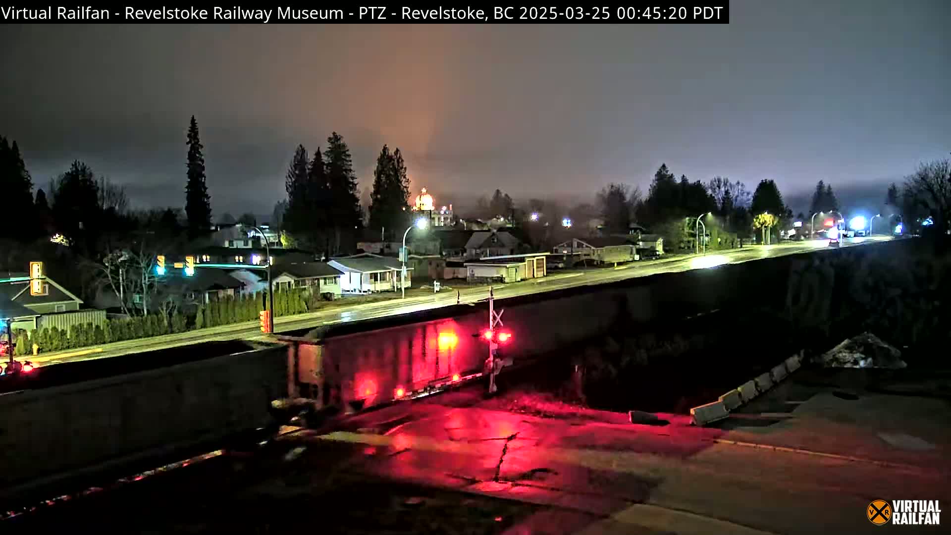 A long freight train with red lights illuminates a rain-slicked road at night, passing in front of a residential area with houses and trees under a hazy sky.