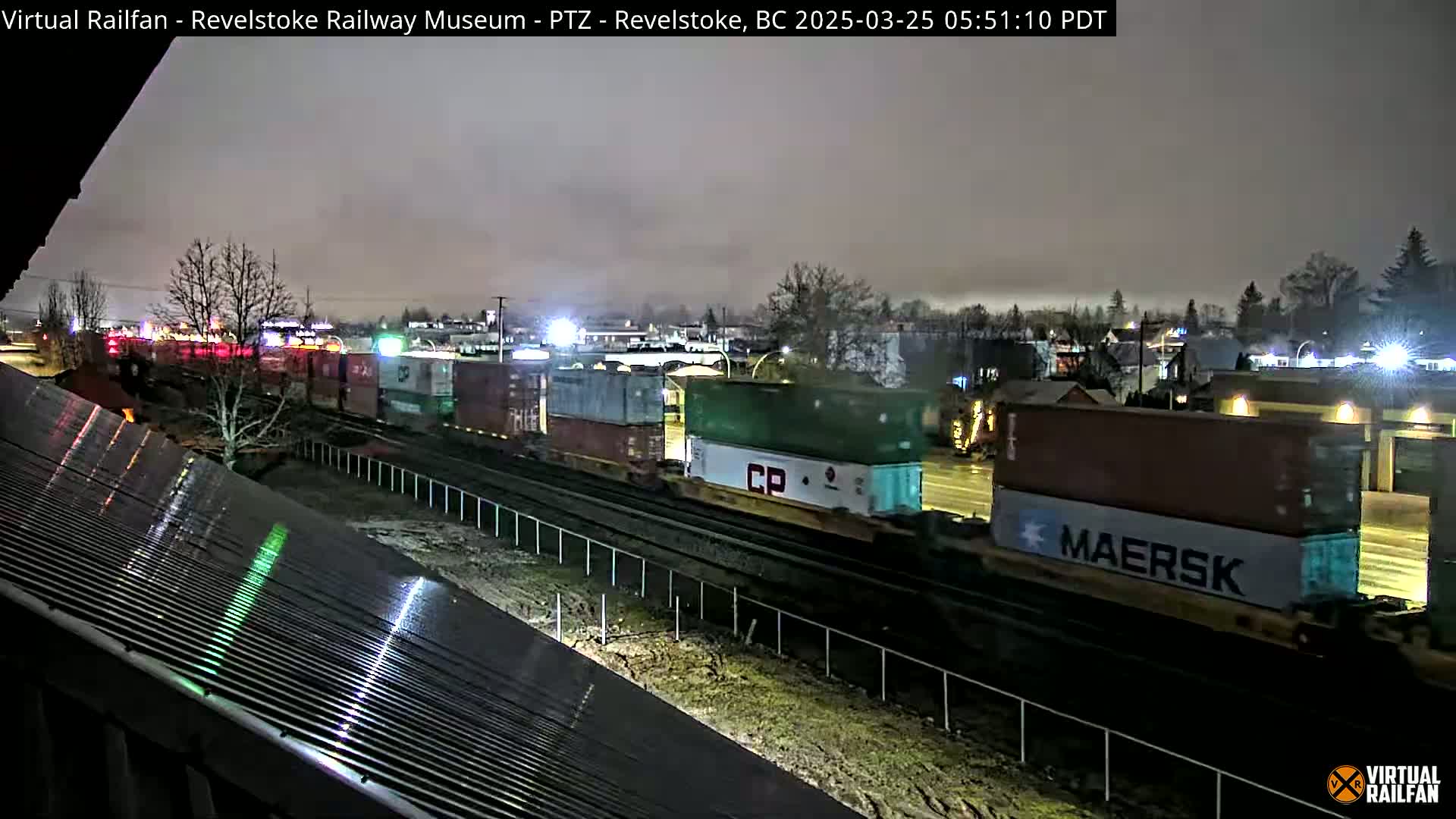 A long freight train carrying numerous shipping containers moves along railroad tracks at night under a cloudy sky.