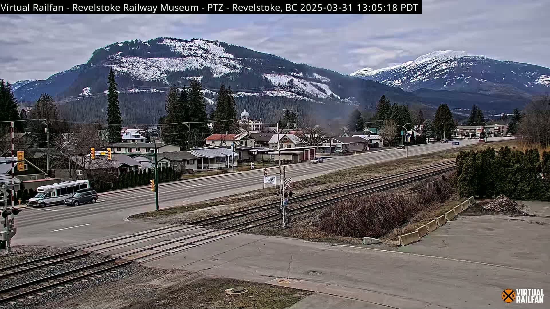A partly cloudy day reveals a snow-dusted mountain range overlooking a town with a roadway and train tracks intersecting at a level crossing.