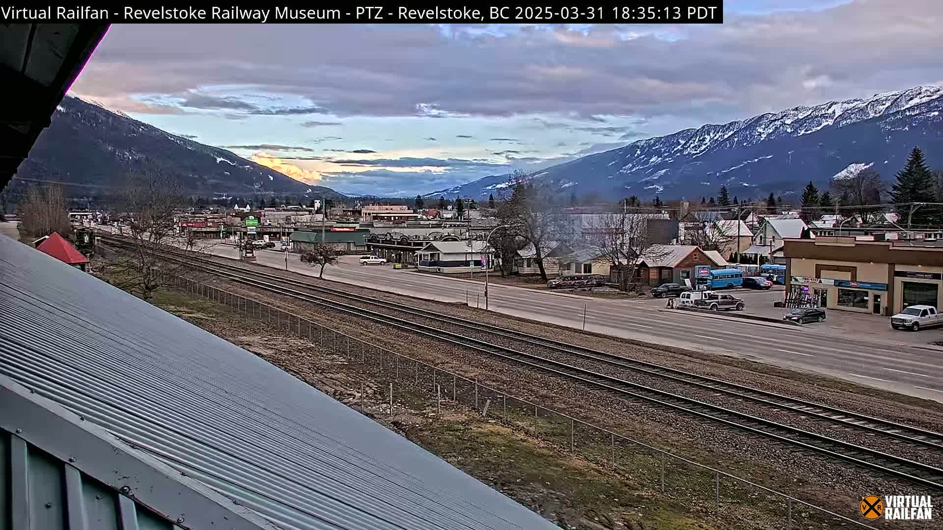 A view of a town nestled in a valley between snow-capped mountains under a partly cloudy sky.