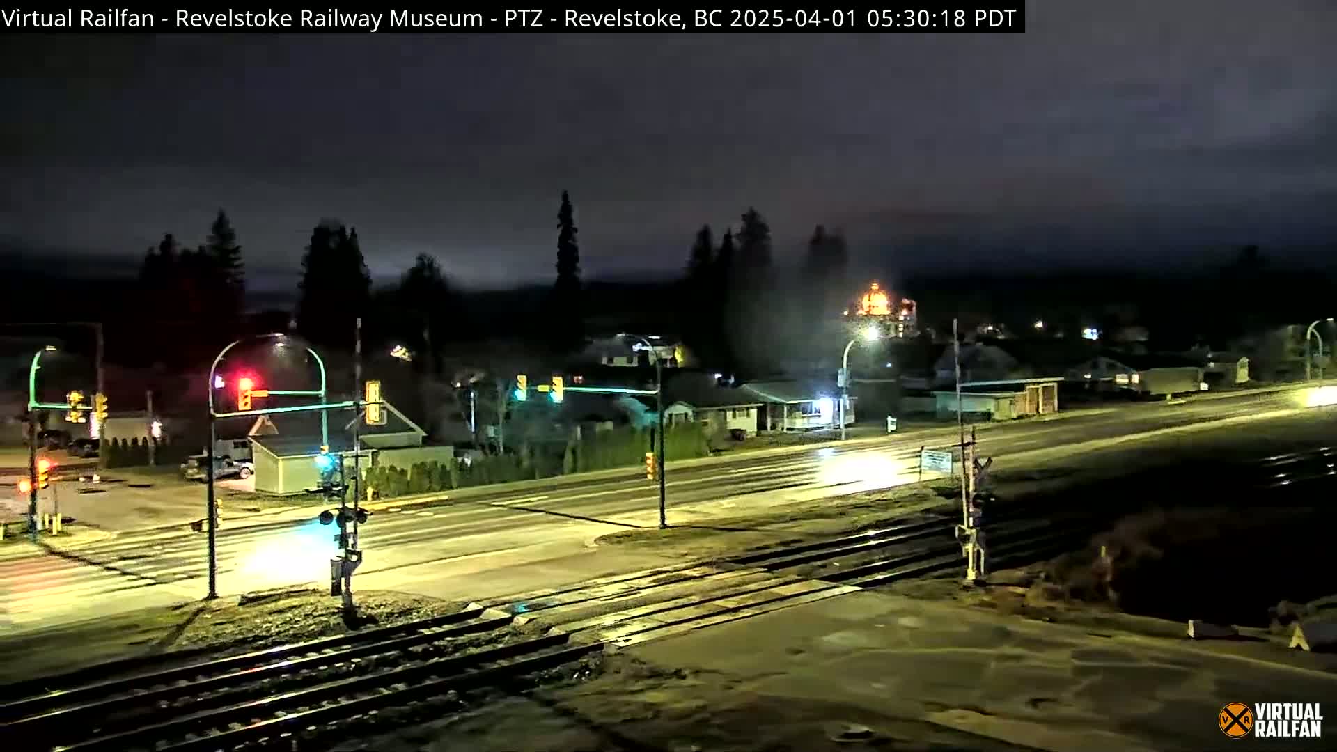 A nighttime view of a wet road intersecting with railroad tracks, passing through a small town under a dark, overcast sky.