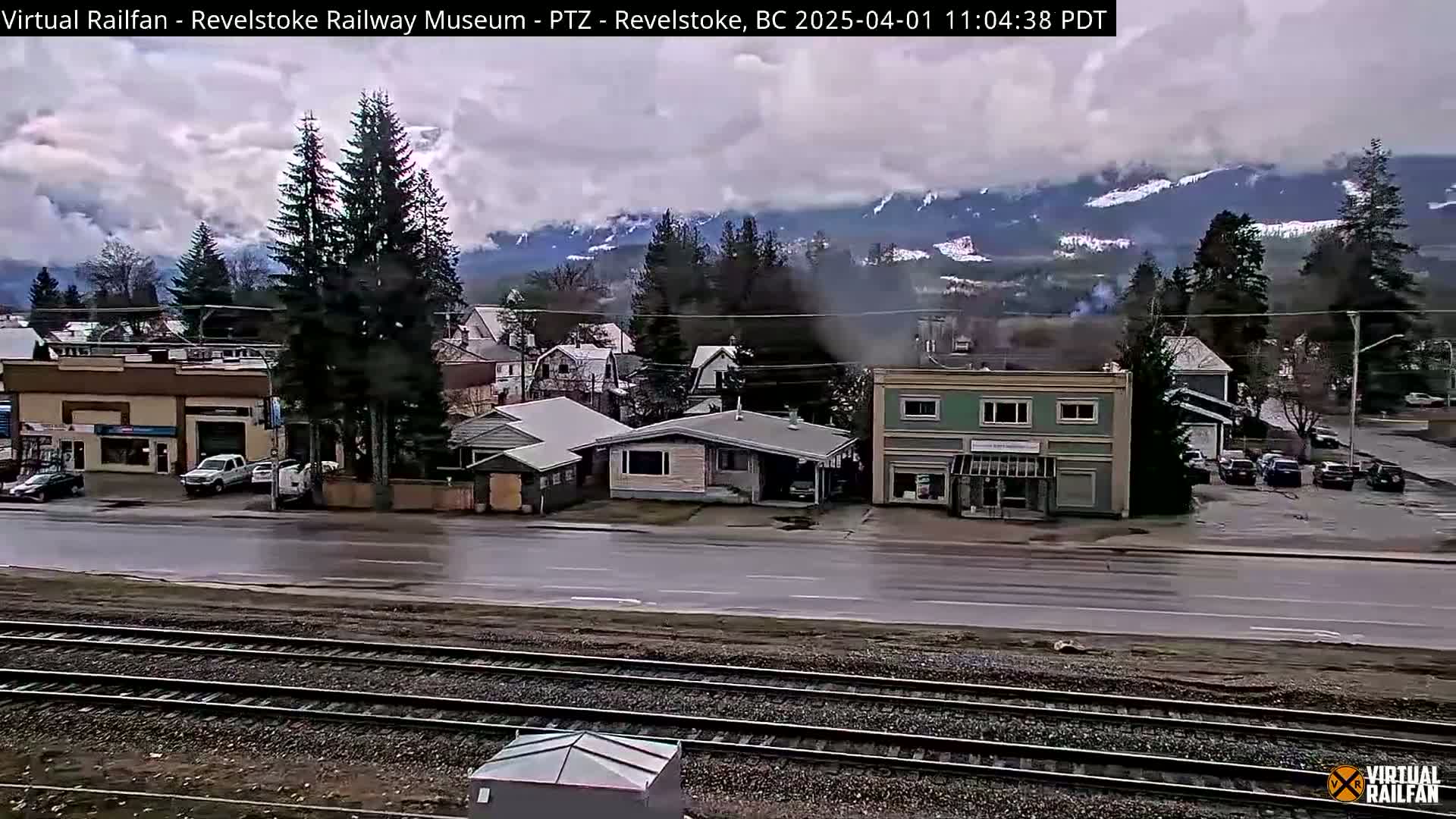 A view of a town with a railway track in the foreground, under overcast skies, with mountains in the background showing some snow.