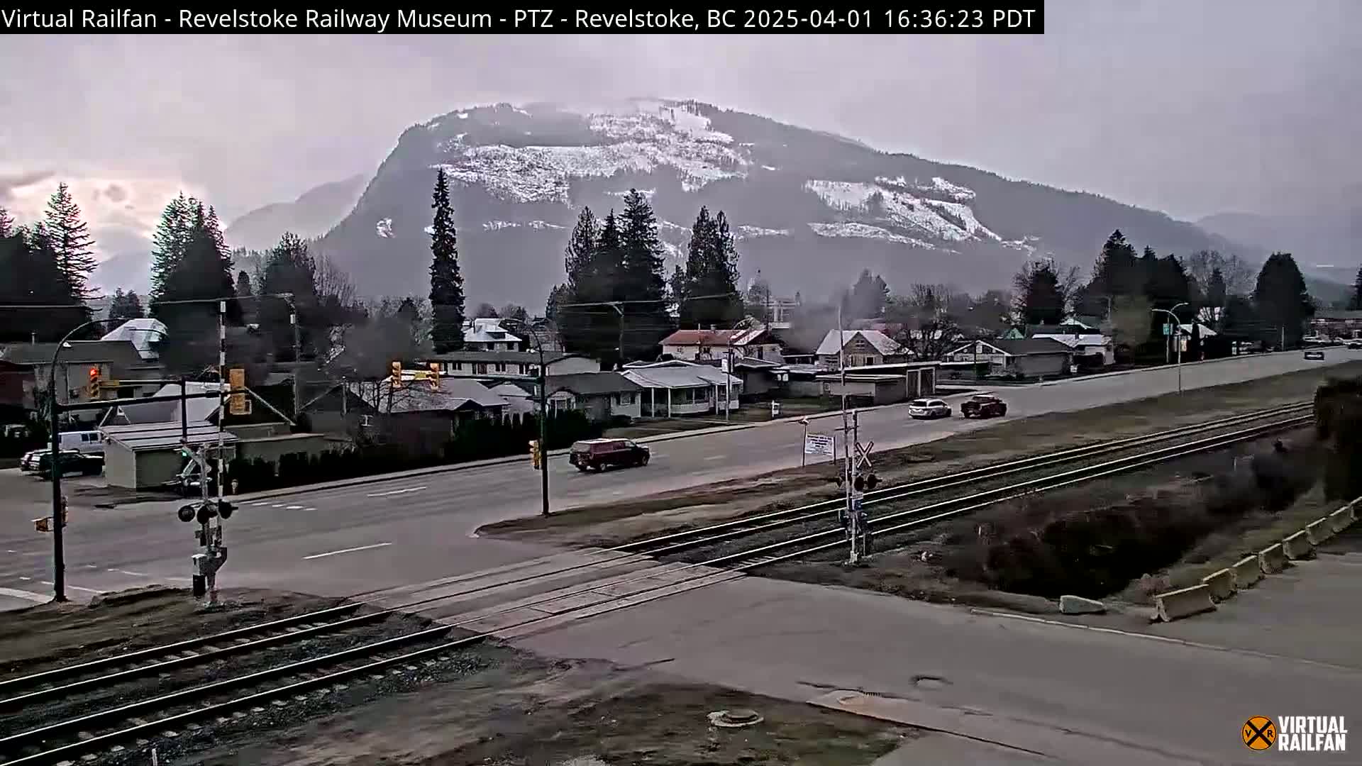 A partly cloudy day reveals a snow-dusted mountain behind a town with a road and railway crossing, with a few vehicles visible.