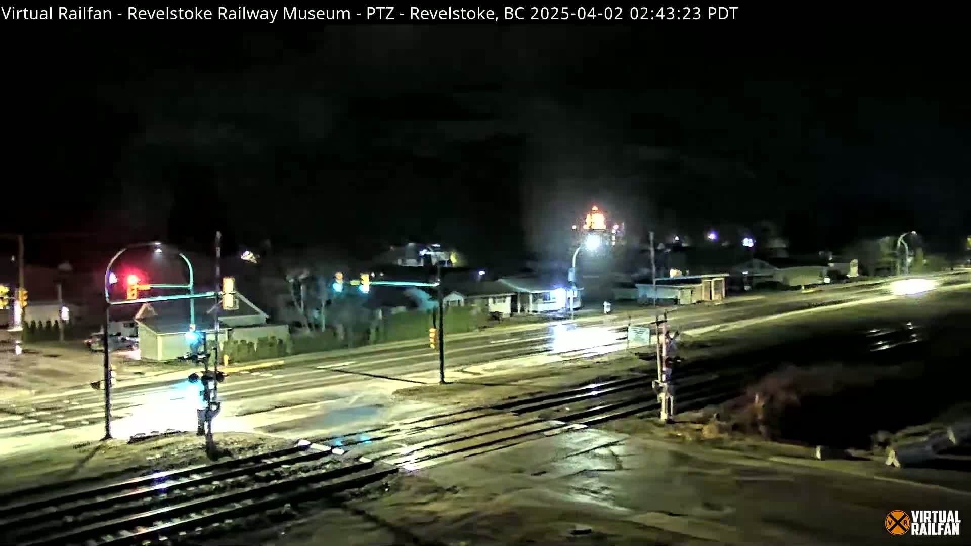 A nighttime view of a rain-slicked road and railroad crossing with traffic lights, streetlights, and buildings in the background.