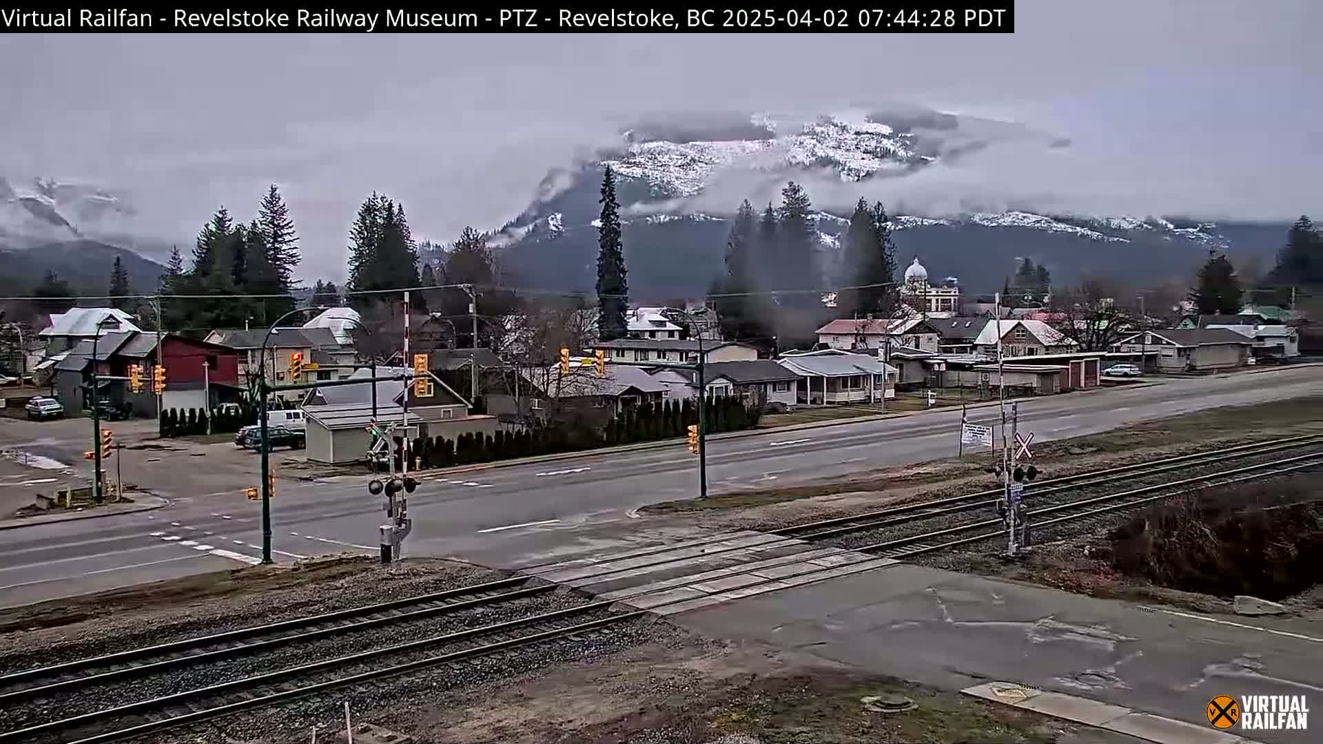 A town with houses and a railway crossing is seen under an overcast sky with snow-capped mountains in the background.